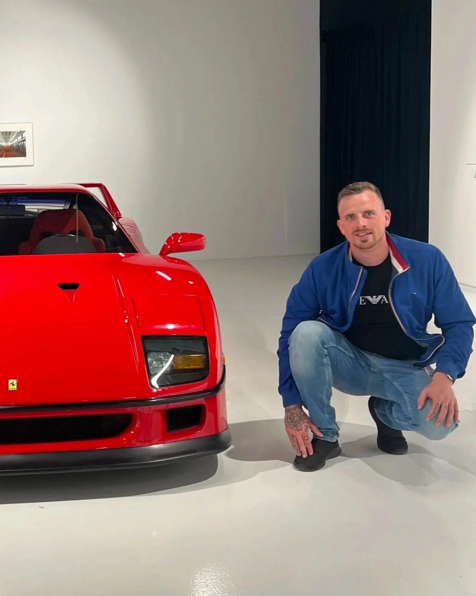 Man met kort haar, baard en tattoos, die in een moderne tentoonstellingsruimte voor een rode Ferrari F40 knielt, met witte muren en een zwarte deur op de achtergrond.