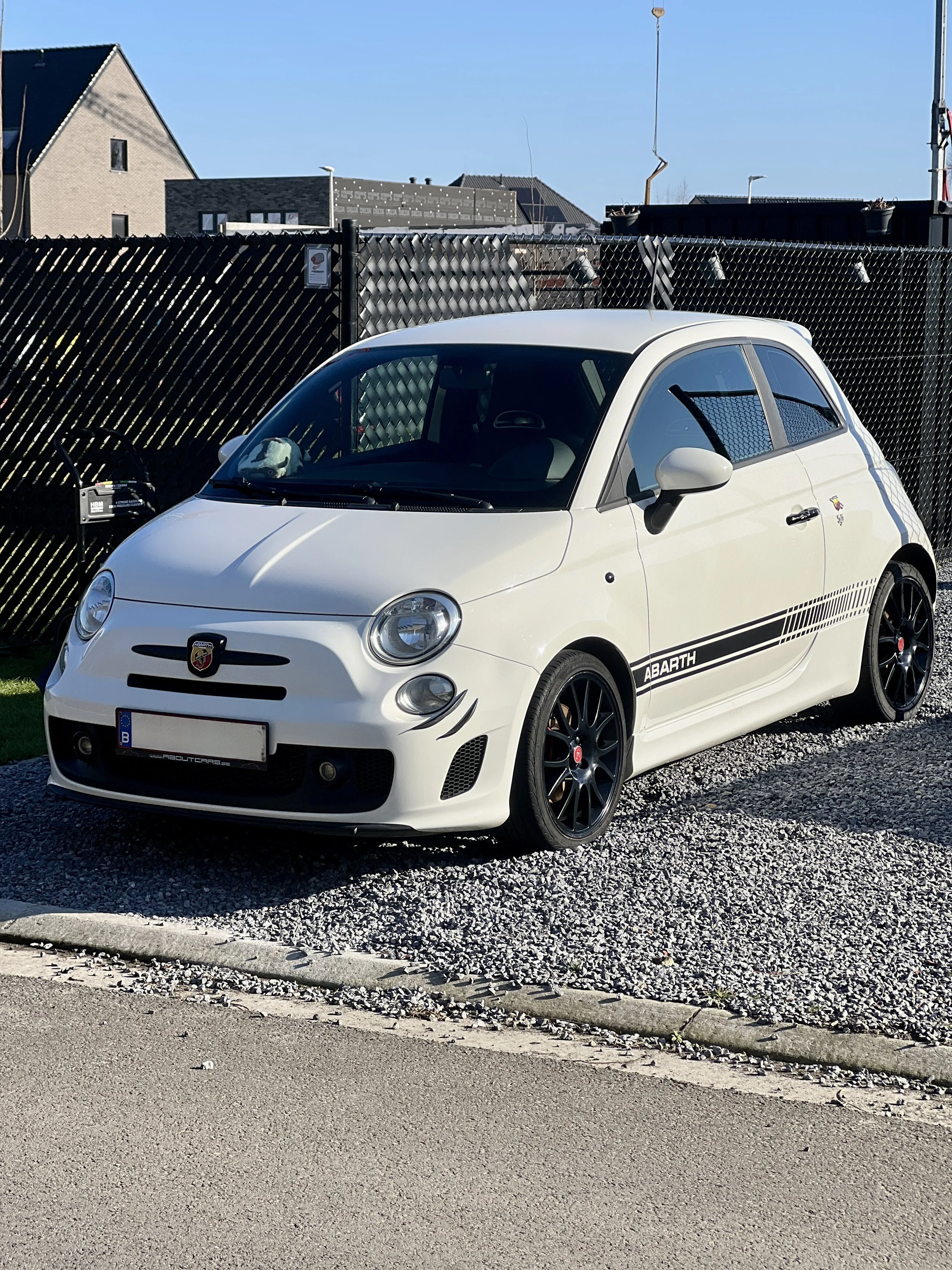 Een witte Fiat Abarth 595 met zwarte velgen geparkeerd op een grindoprit, met een zwarte hek en op de achtergrond een heldere blauwe hemel.