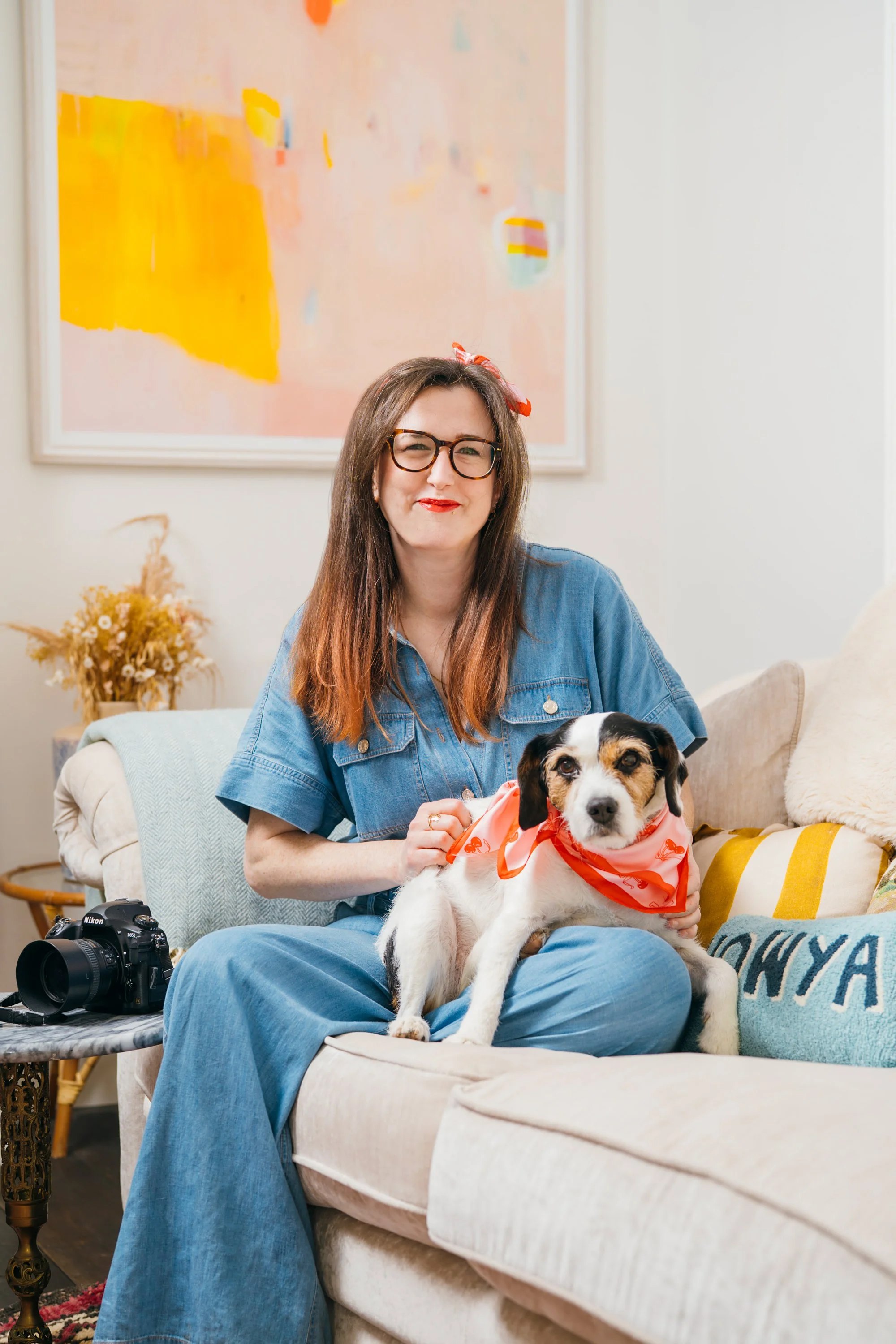 El Keegan with glasses and long brown hair sitting on a beige couch, holding Fuji, a black and white dog wearing a red bandana, in a cozy living room with a colourful abstract painting and a camera on a side table.