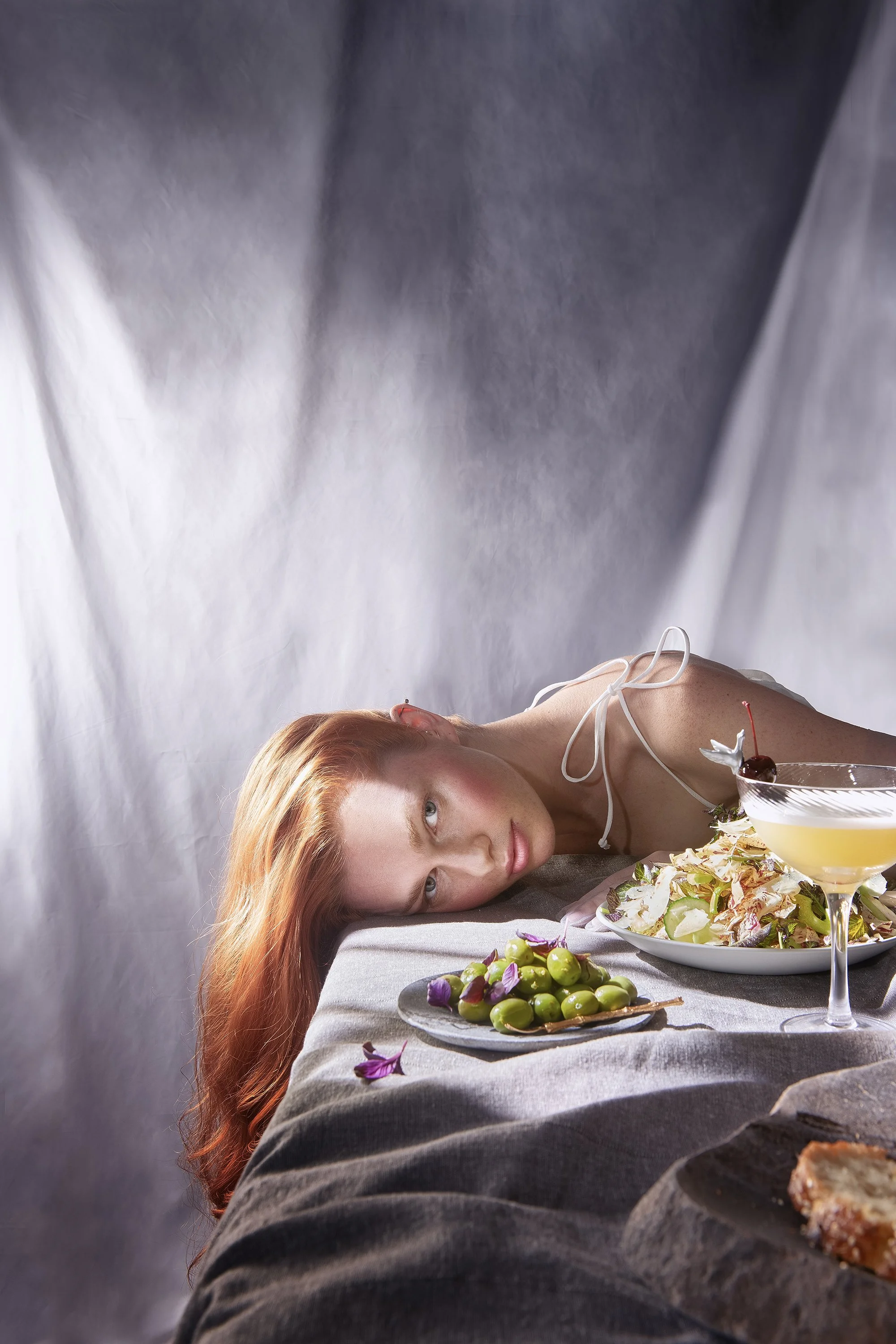   A model's hair flows off the side of a table with a salad, olives and a cocktail. She looks passed the food to the camera. Photo created by Dublin Photographer El Keegan, Prop Styling by Charlotte Carson, Food styling by Sage Dakota, Hair and Makeu