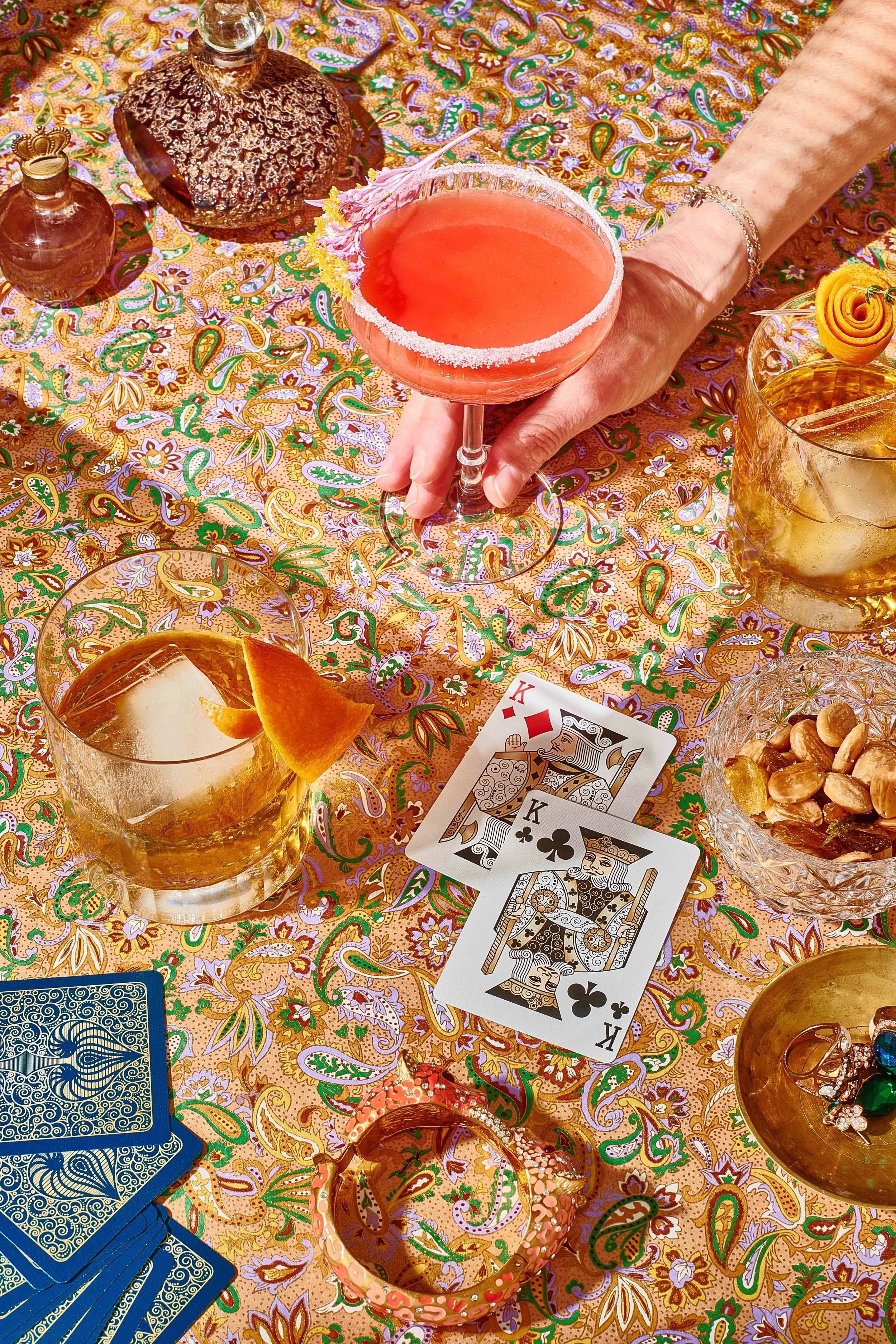   A woman's hand holding a tall stem coral cocktail on colourful wallpaper with another whiskey based cocktail and some playing cards. Photography and Retouching by El Keegan Dublin Ireland  

