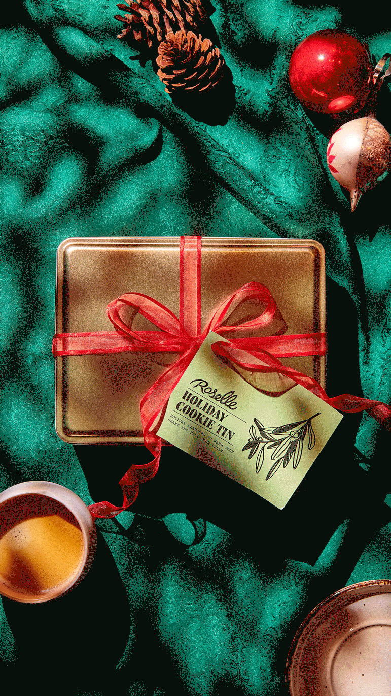   A gif of a Roselle holiday biscuit tin being eaten. Shortbread, gingerbread, cocoa cookies and biscotti are stacked in a gold tin wrapped in a red ribbon. A coffee, plate and Christmas decorations on placed on the deep green tablecloth.  Created by