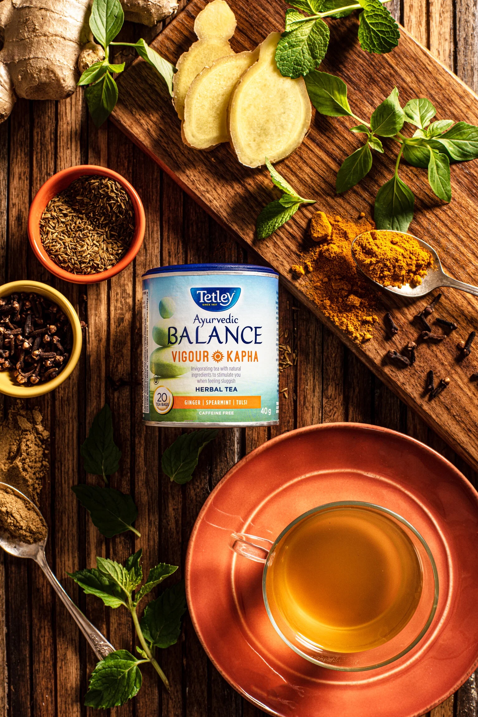   A rustic overhead photo of Tetley tea surrounded by spices, including, turmeric, cumin seeds, and cloves. Sliced ginger and herbs are laid out on a wooden cutting board and a cup of tea in a glass mug is placed on a side plate. Commercial photograp