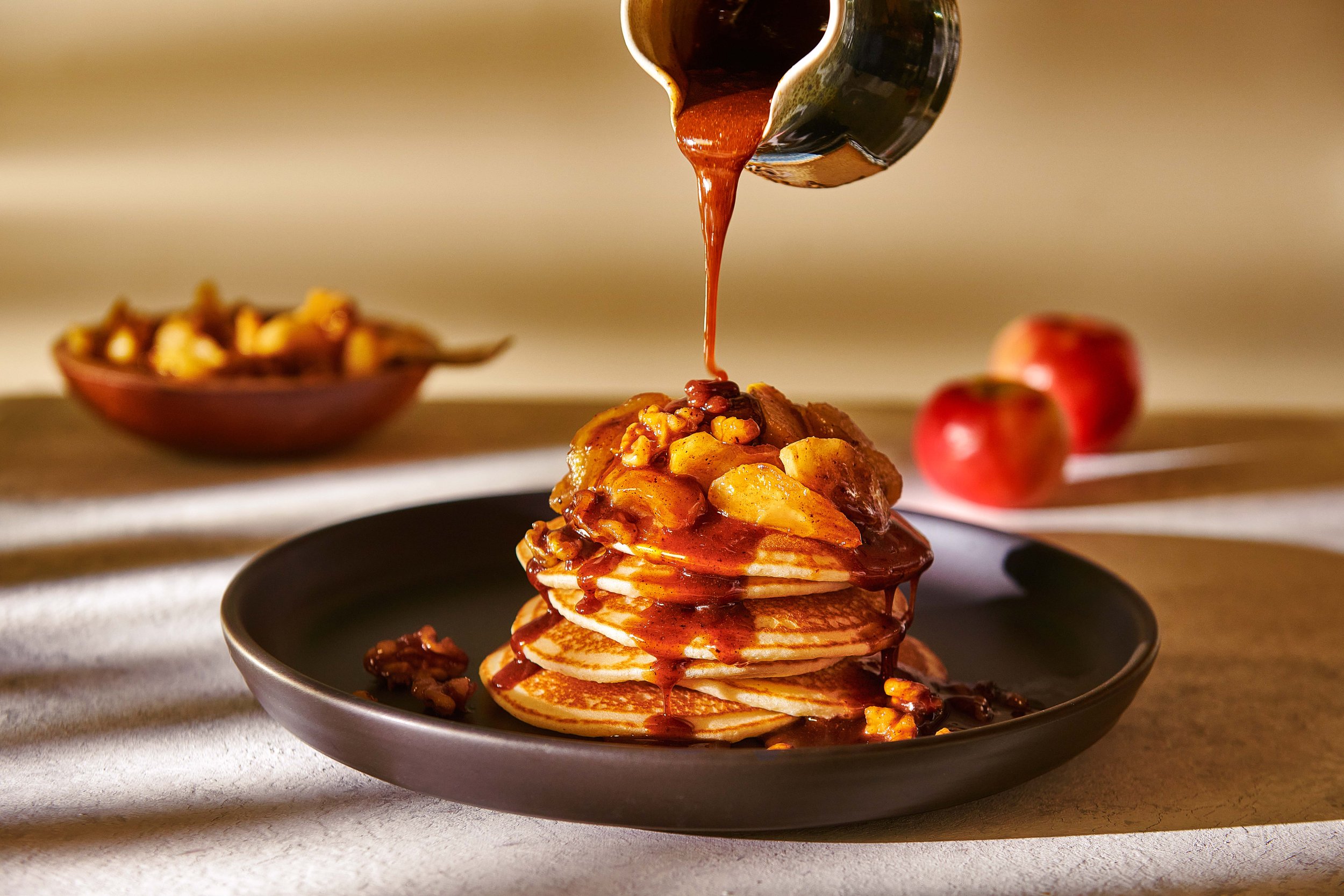  Maple syrup pours over a stack of thick, fluffy pancakes with sliced, stewed apple. Food photography by El Keegan, Dublin, Ireland.  