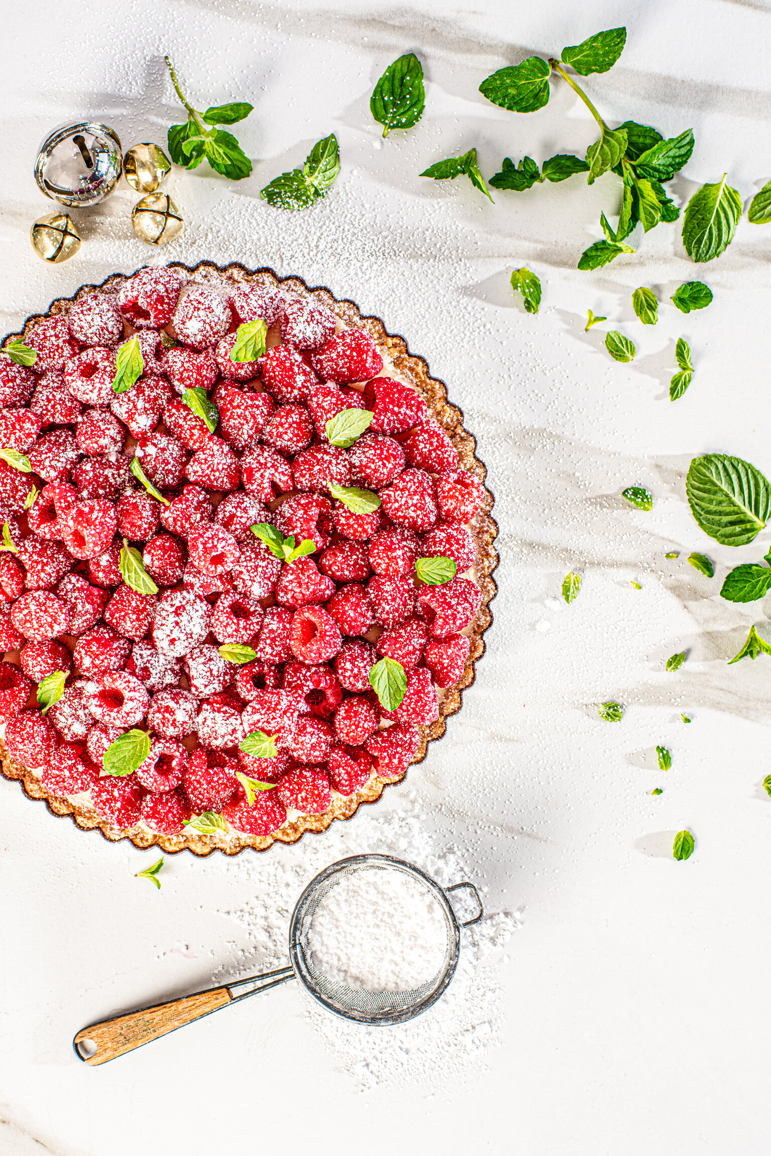  A fresh raspberry tart sits on a marble tabletop with a sieve nearby containing sprinkled sugar. Photographed by Dublin Food Photographer, El Keegan.  