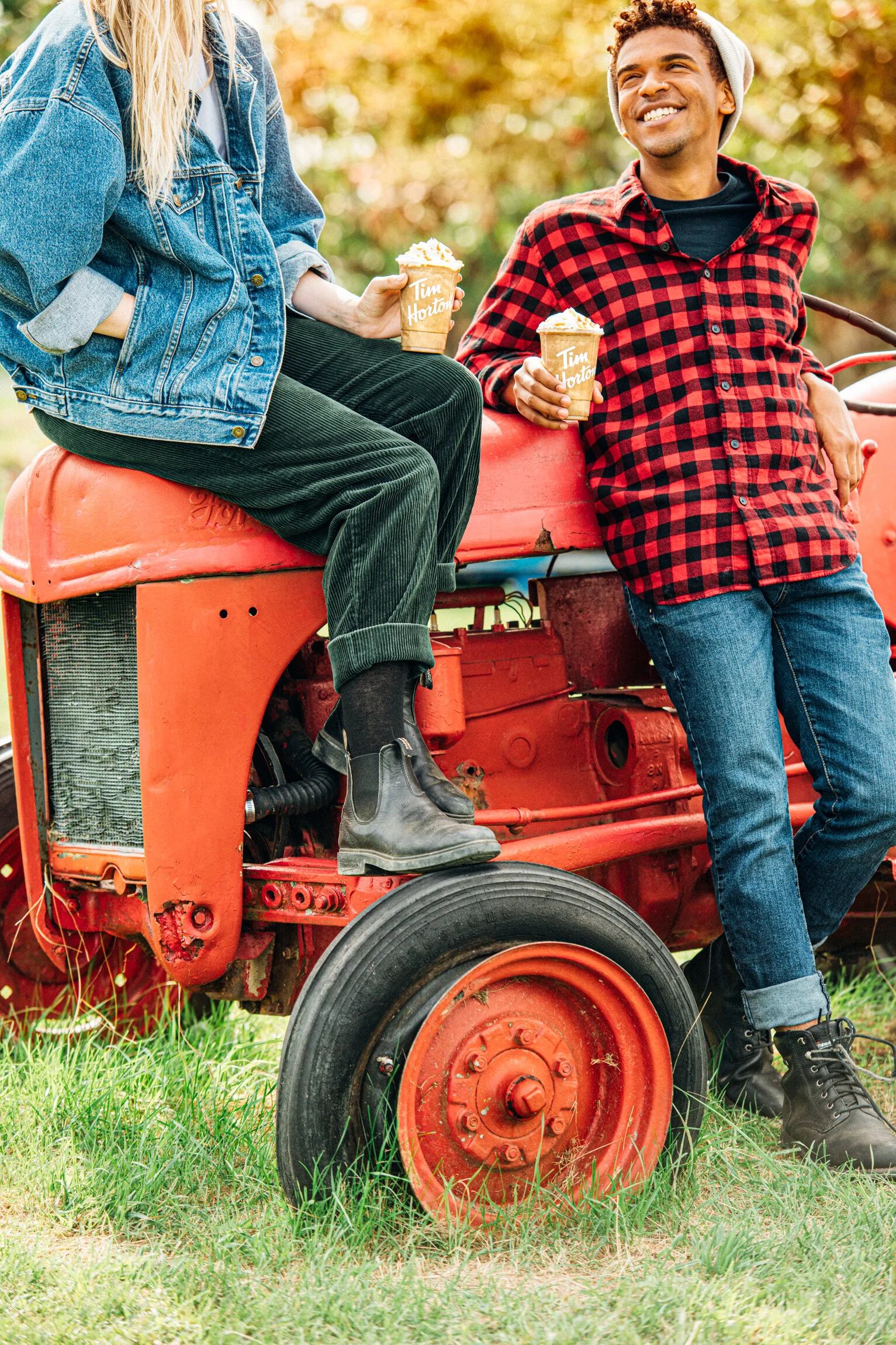   A young couple lean against a red tractor enjoying Tim Hortons iced coffees. A man in a red and black buffalo plaid shirt, blue jeans and black boots looks up smiling at a woman in a denim jacket. Lifestyle photography created by Dublin Photographe