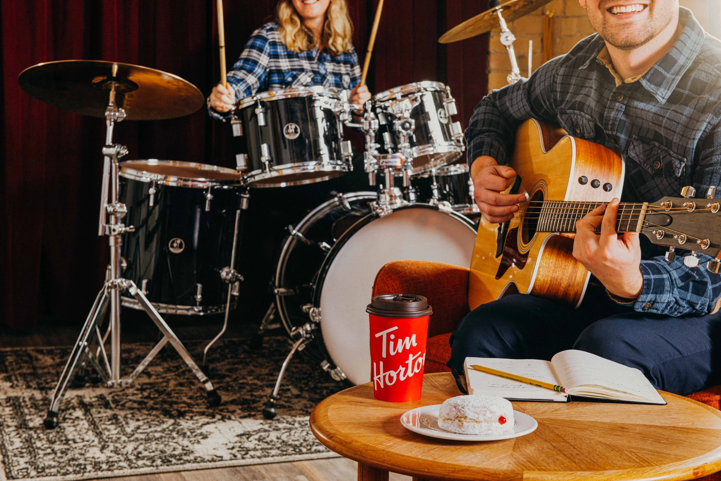   A smiling man plays guitar at a coffee table with a notebook, pencil, a cup of Tim Hortons coffee and a powdered jam donut. A woman plays the drums in the background. Jam session photography and retouching by El Keegan, Dublin, for Tim Hortons.  