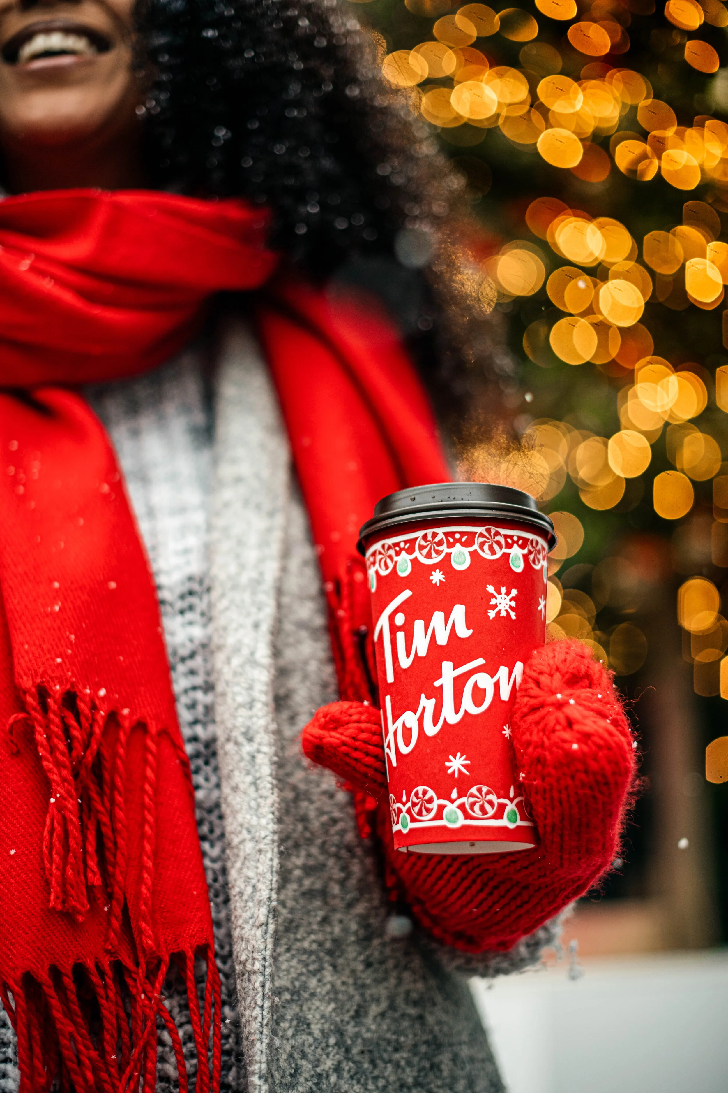  A smiling woman wrapped up in a red scar and mittens holds a cup of Tim Hortons coffee in front of a wall of Christmas lights. Drinks photography by Dublin Photographer, El Keegan.  