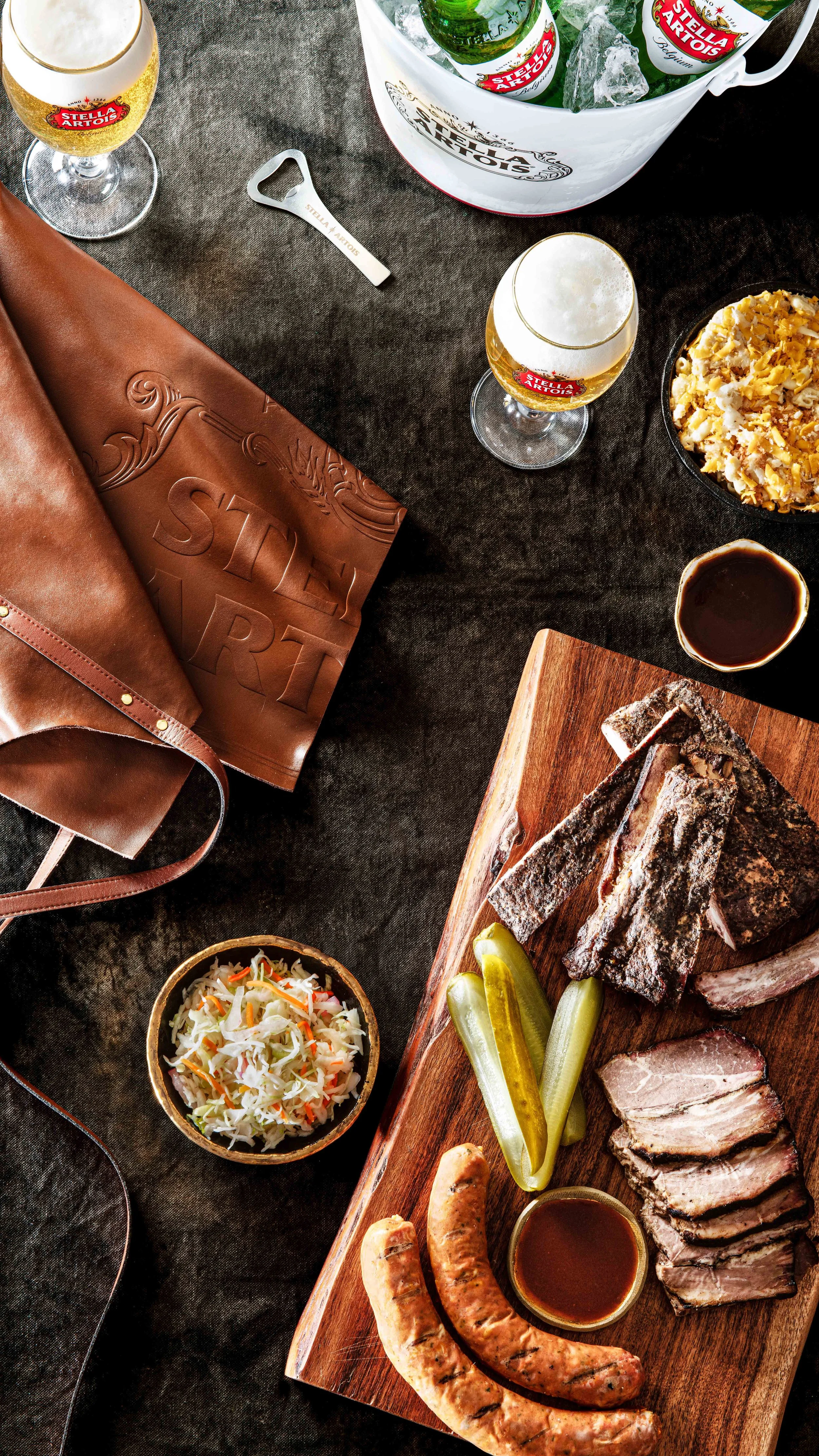   Overhead image of a barbecue spread of ribs, sausages, coleslaw, pork and pickles on a chopping board. Two Stella Artois beers sit in stemmed glasses, an apron and a bottle opener lie beside an ice bucket holding beer bottles. Commercial Photograph