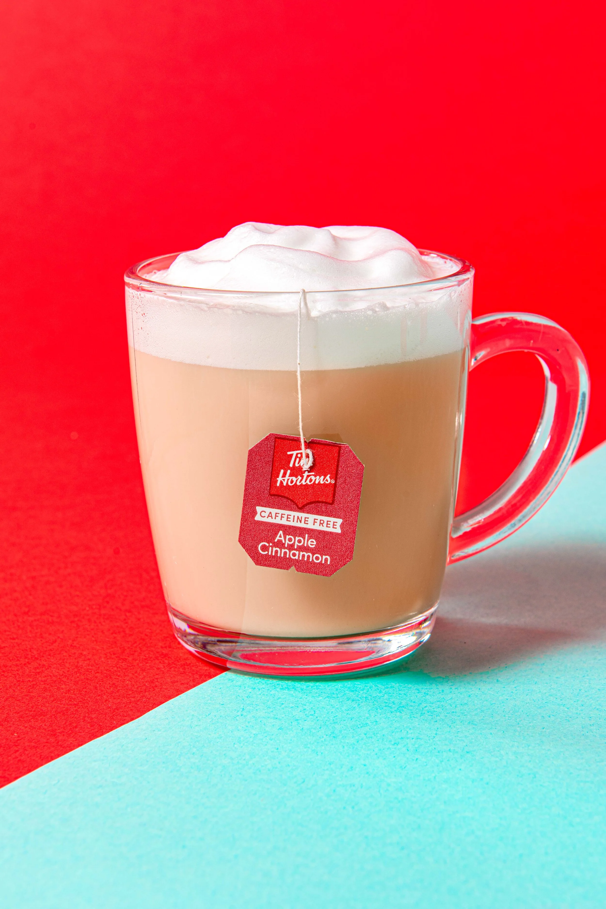  A hot tea latte drink with foamed milk and the tag hanging on the front. Photographed on bright red and blue backgrounds in a glass mug. Image created by El Keegan for Tim Hortons. 