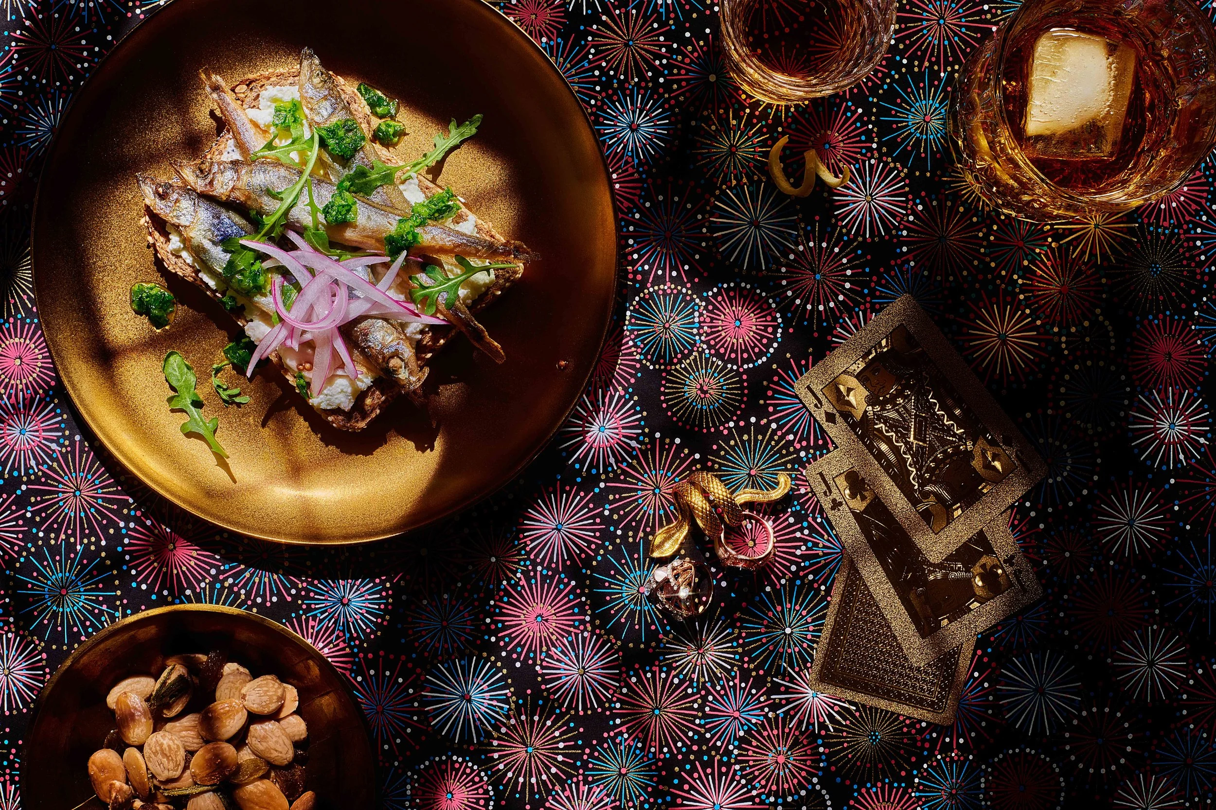  An overhead image of a gold plate with fish and sliced onions. Two tumblers of whiskey sit in the corner and playing cards sit on the patterned surface. Food photography by El Keegan, Dublin.  