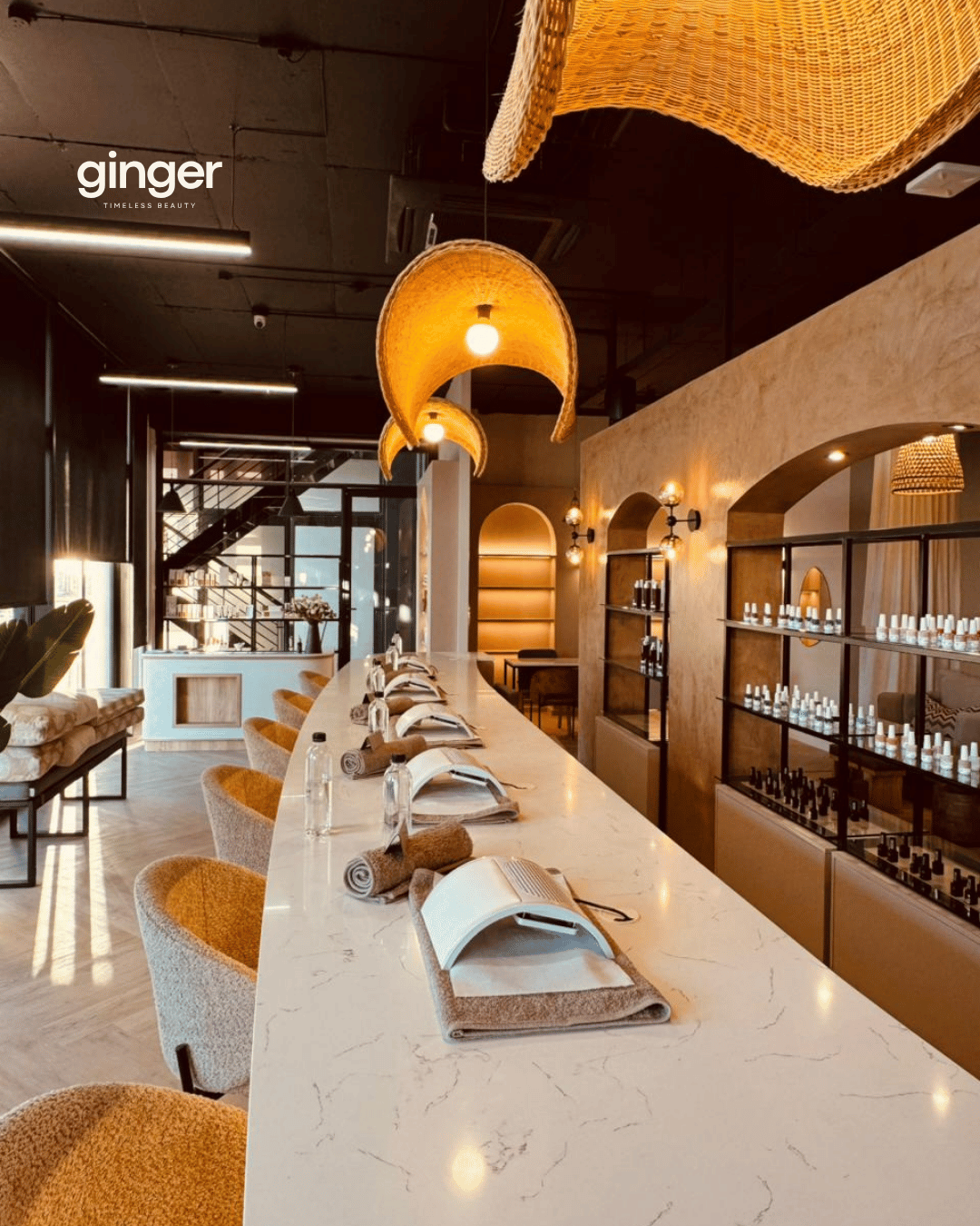 Interior of a modern beauty salon or spa with a long white marble counter, chairs, reading materials, rolled towels, and bottles of nail polish , with warm lighting and decorative lighting fixtures. The image promotes client Ginger Spa
