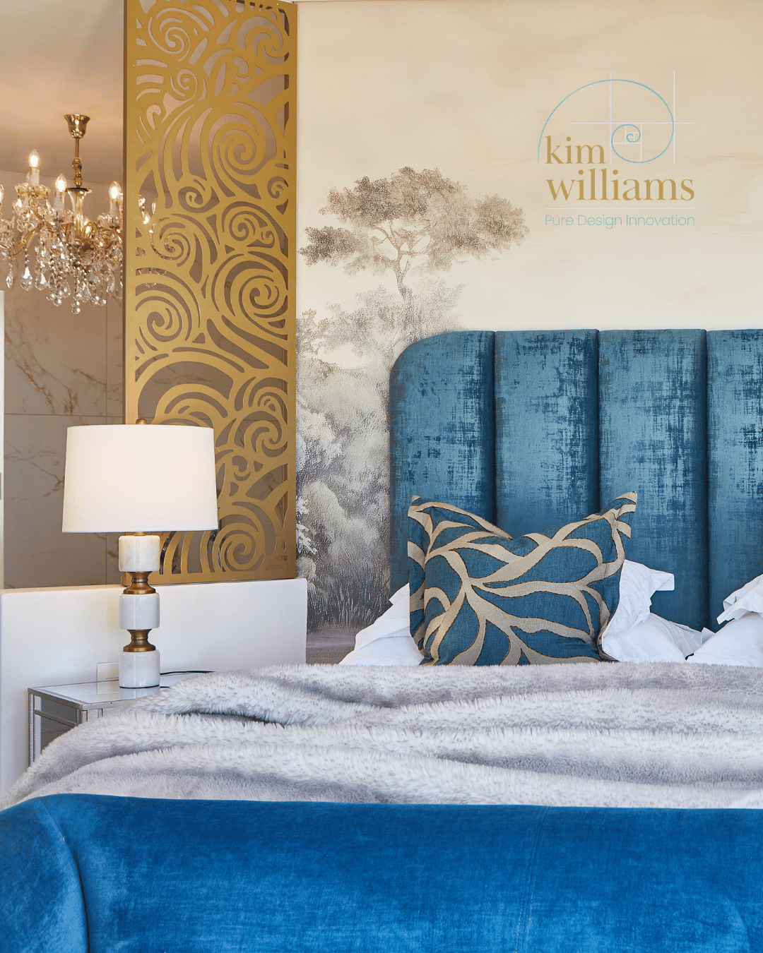 A bedroom with a blue upholstered headboard, decorative pillow, white bedding, and a white lampshade on a side table. The wall features a mural of a tree and a logo reading 'kim williams, Pure Design Innovation', with a decorative gold panel and chandelier in the background.