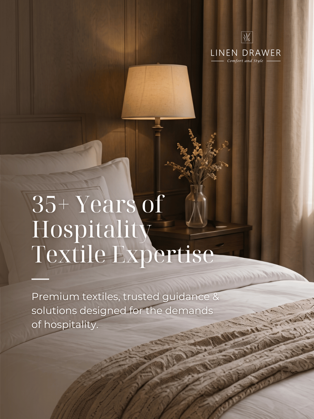 A cozy hotel bedroom with a neatly made bed, a bedside table with a table lamp, a vase with dried flowers, and curtains on a window. The image promotes linen drawer's experience in hospitality textiles.
