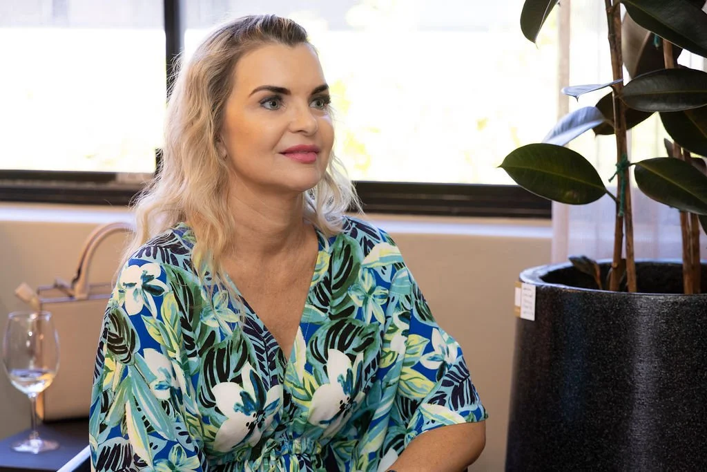 A woman with wavy blonde hair wearing a colorful floral dress sitting indoors by a window, next to a large potted plant. Erica Wagner the founder of birdseyedigital.co.za