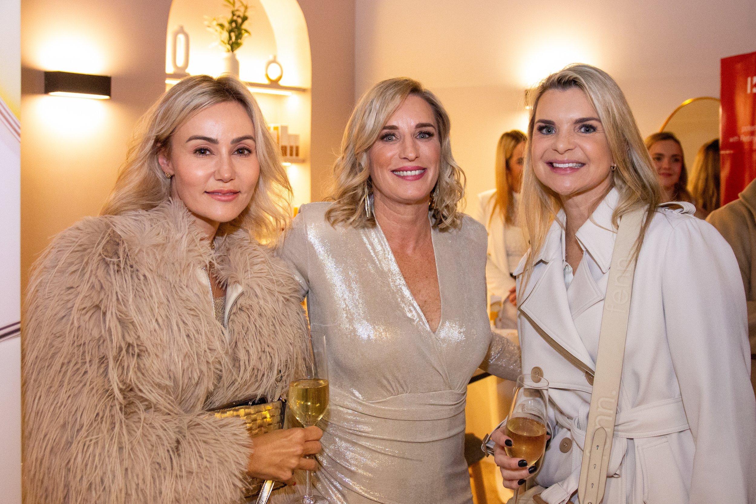 Three women with blonde hair dressed elegantly, holding glasses of champagne, standing close together and smiling at a social gathering. Including the business owner Erica Wagner and a Client Tanya from Ginger Spa