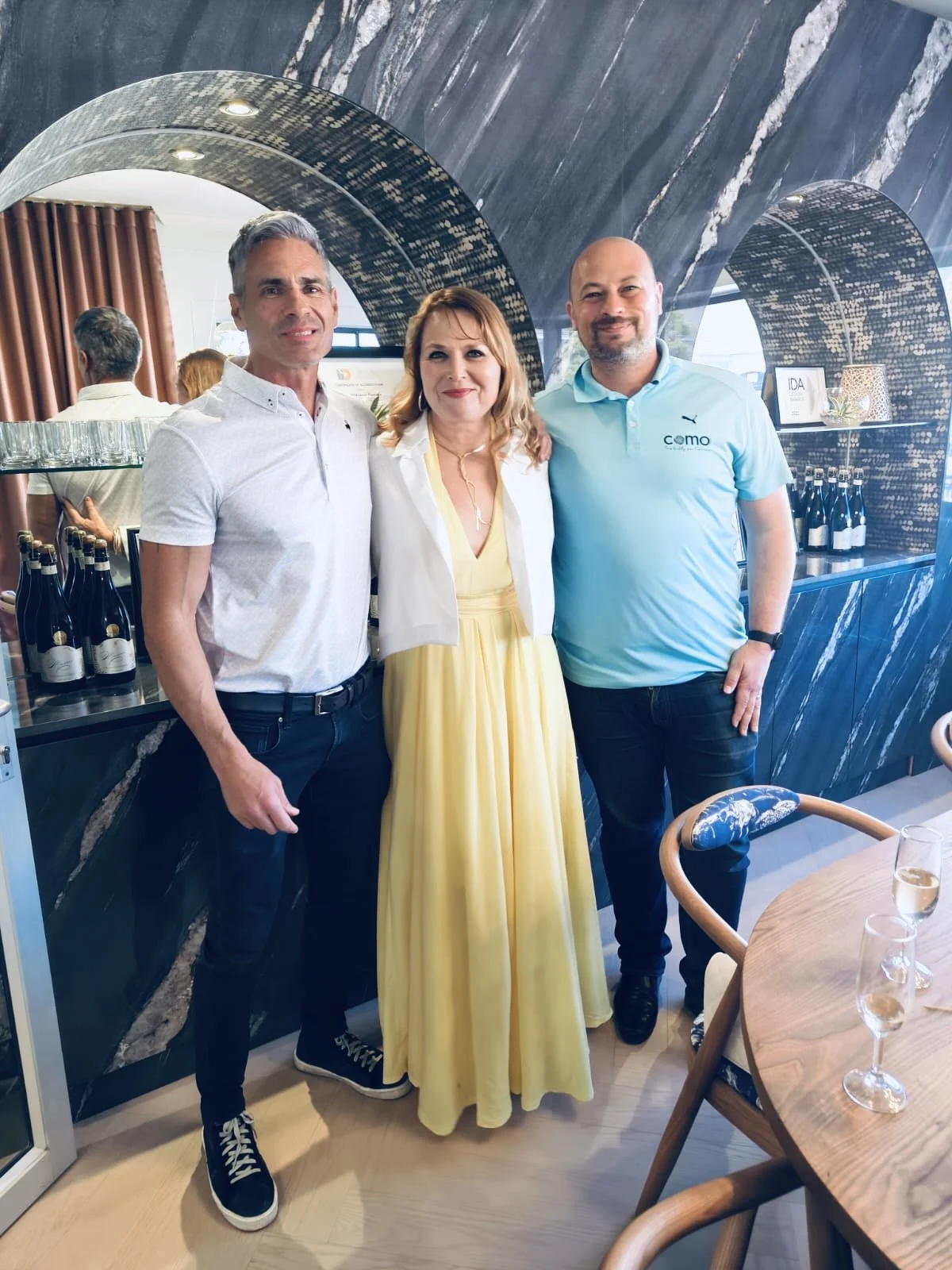 Three people standing together in a restaurant or cafe. Two men and one woman, all smiling. The man on the left has gray hair, wearing a white shirt and dark pants. The woman in the middle has shoulder-length blonde hair, wearing a white jacket over 