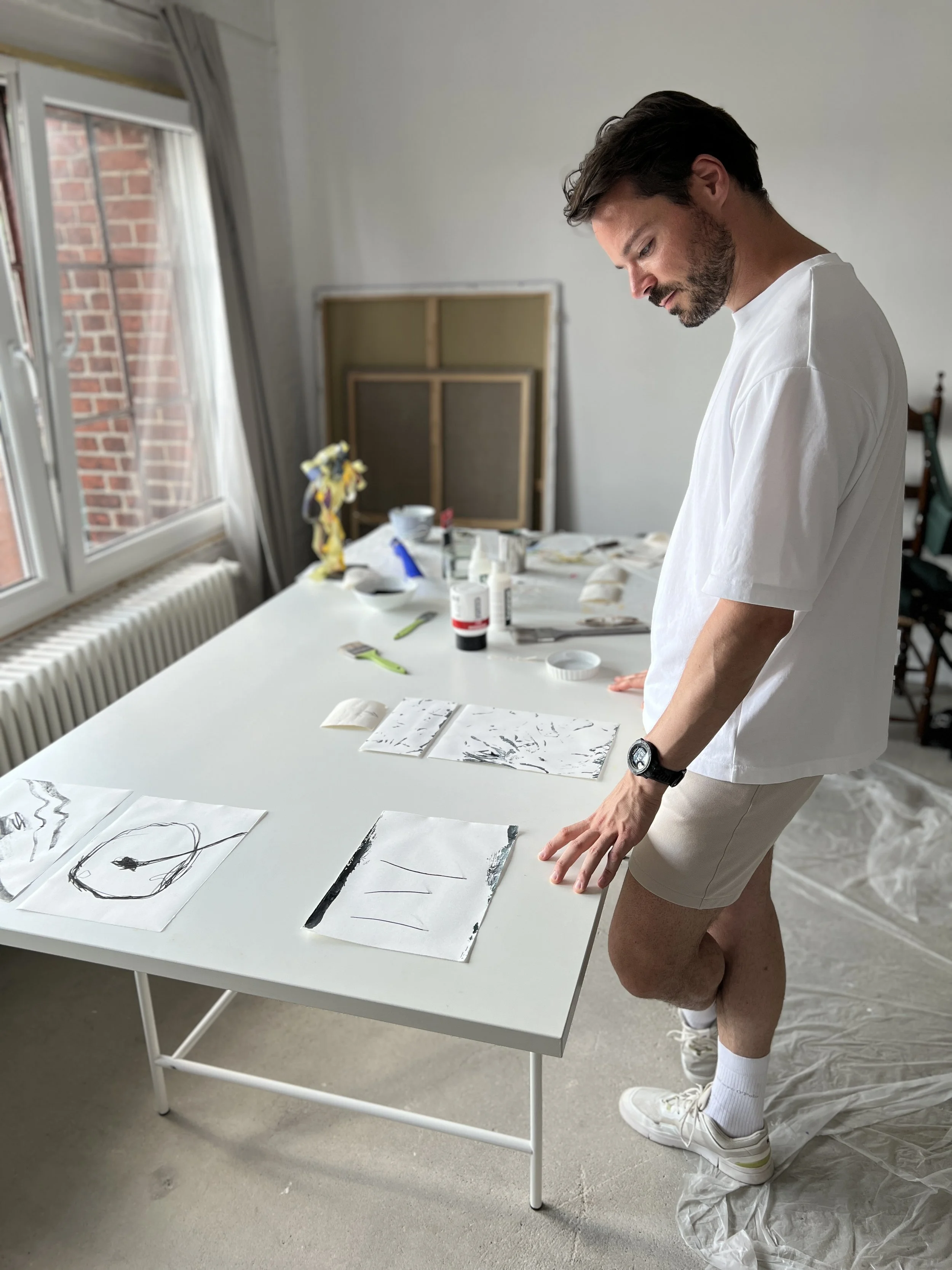 A man with dark hair and a beard wearing a white t-shirt, tan shorts, white sneakers, white socks, and a watch, standing at a white table covered with drawings, art supplies, and paintings, in a room with a window, gray curtains, a radiator, and an art canvas.