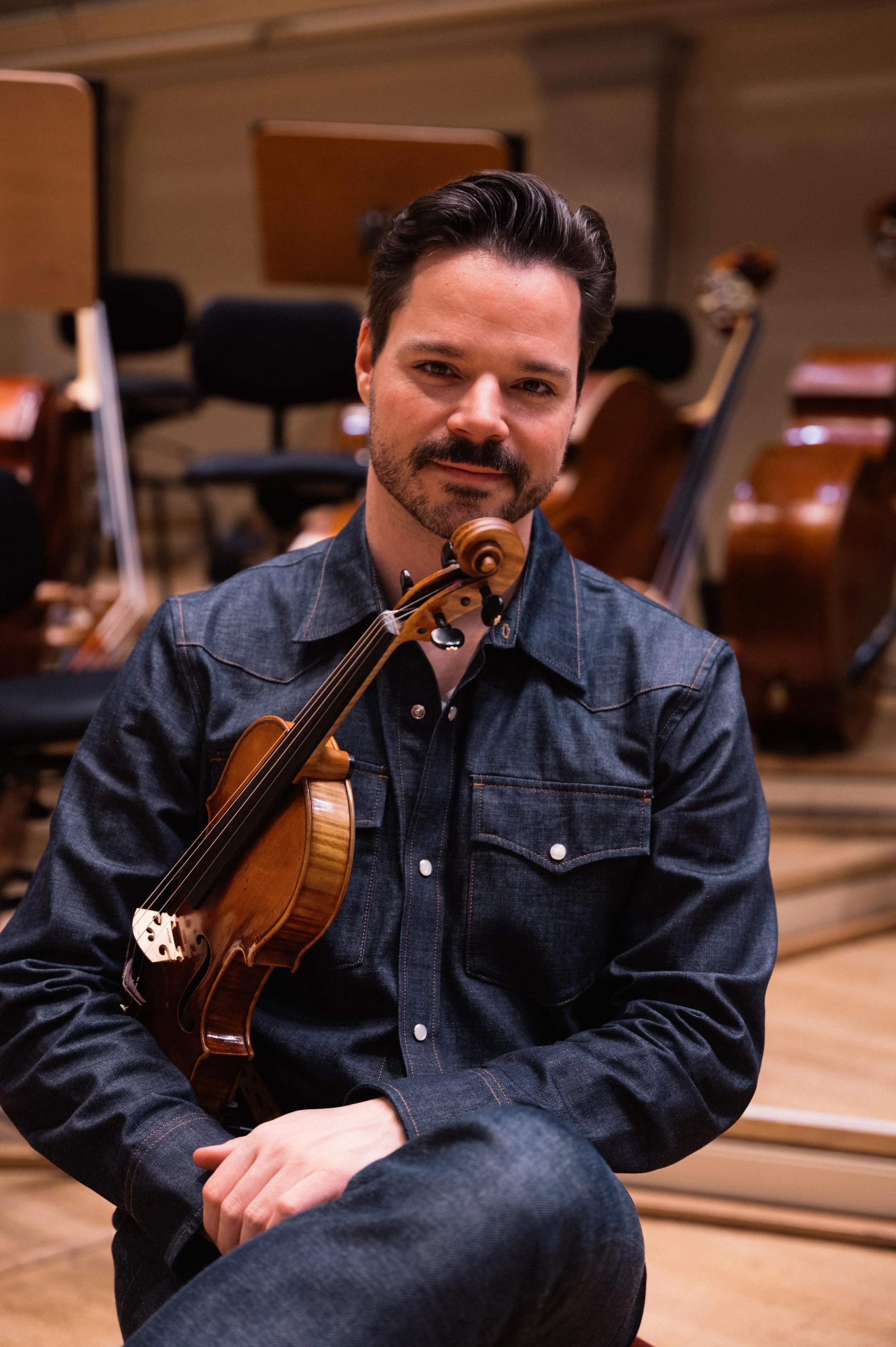A man with dark hair and a beard, wearing a denim shirt, holding a violin, sitting in an orchestra rehearsal room with wooden chairs and music stands.