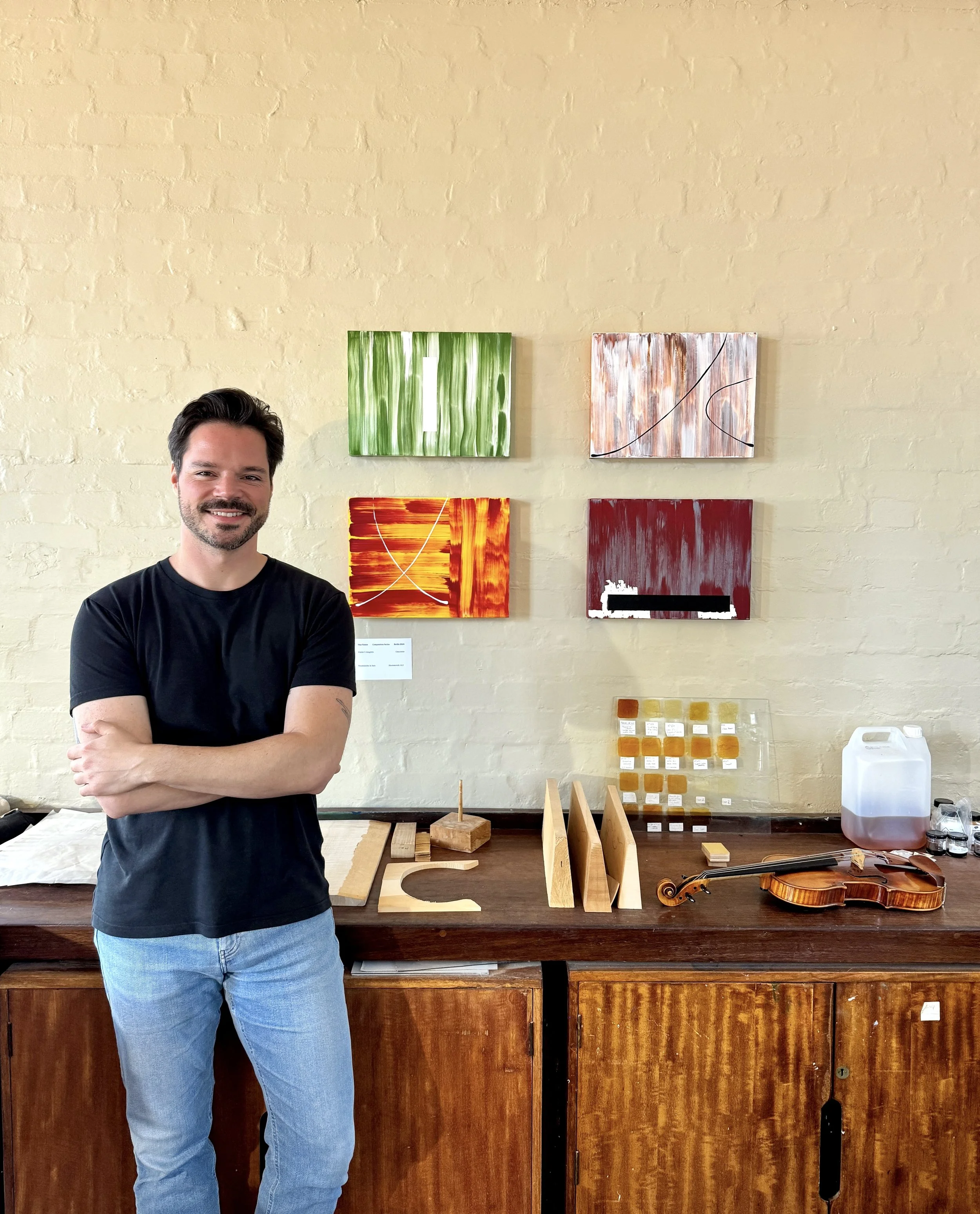 A man with dark hair and a beard smiling, wearing a black T-shirt and blue jeans, standing in front of a wall with colorful abstract paintings. The paintings are rectangular and feature various patterns and colors, with one in green, another in white and orange, and others with red and white. There is a wooden table with various objects, including a violin and sample cards, in front of the man.