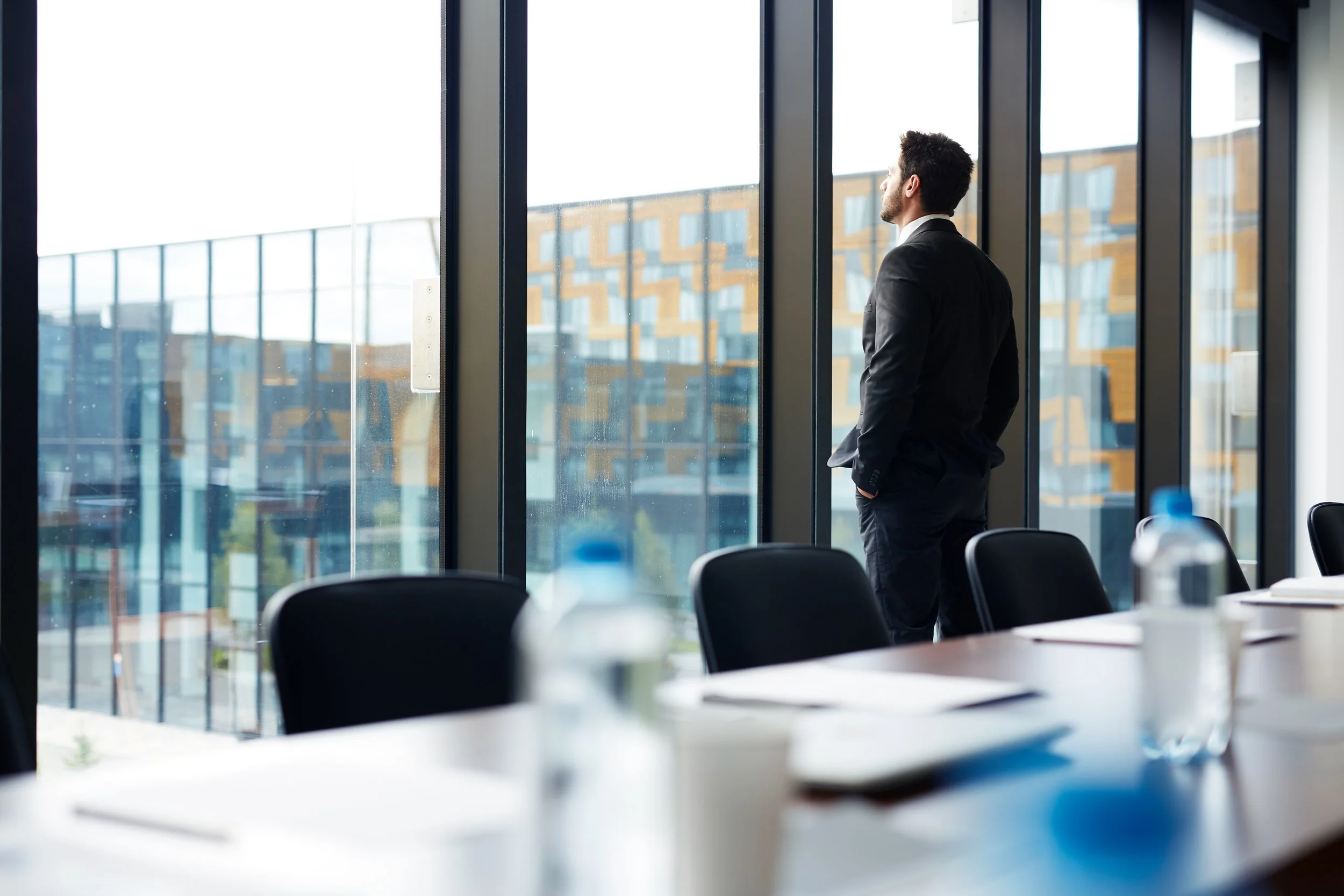 A man in a suit stands by large glass windows, looking outside in a modern office or conference room.