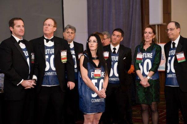 Group of people at a formal event, wearing blue shirts with campaign slogans, standing together indoors.