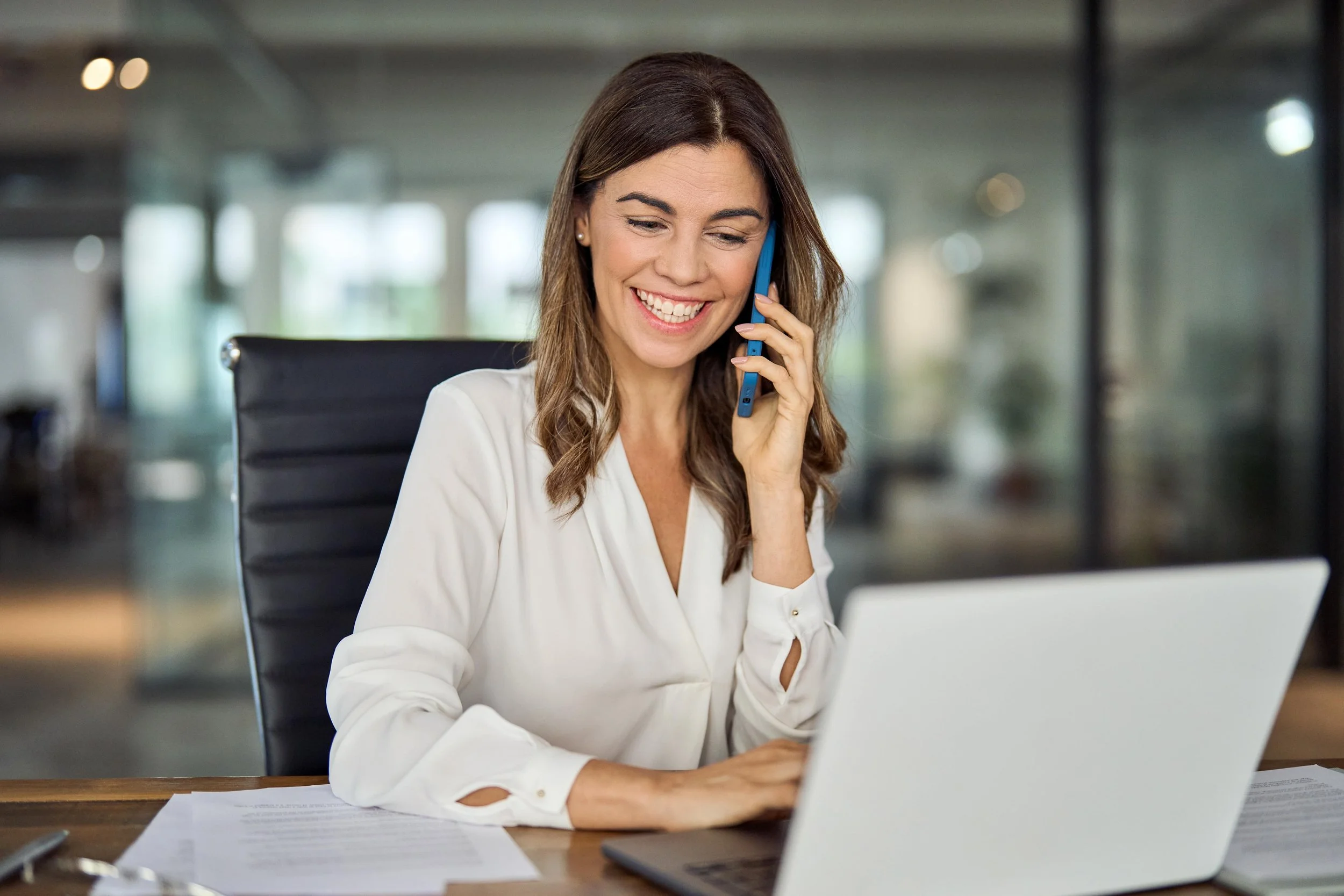 A woman with long brown hair, wearing a white blouse, smiling and talking on a blue mobile phone while sitting at a desk with papers and a laptop in a modern office environment.