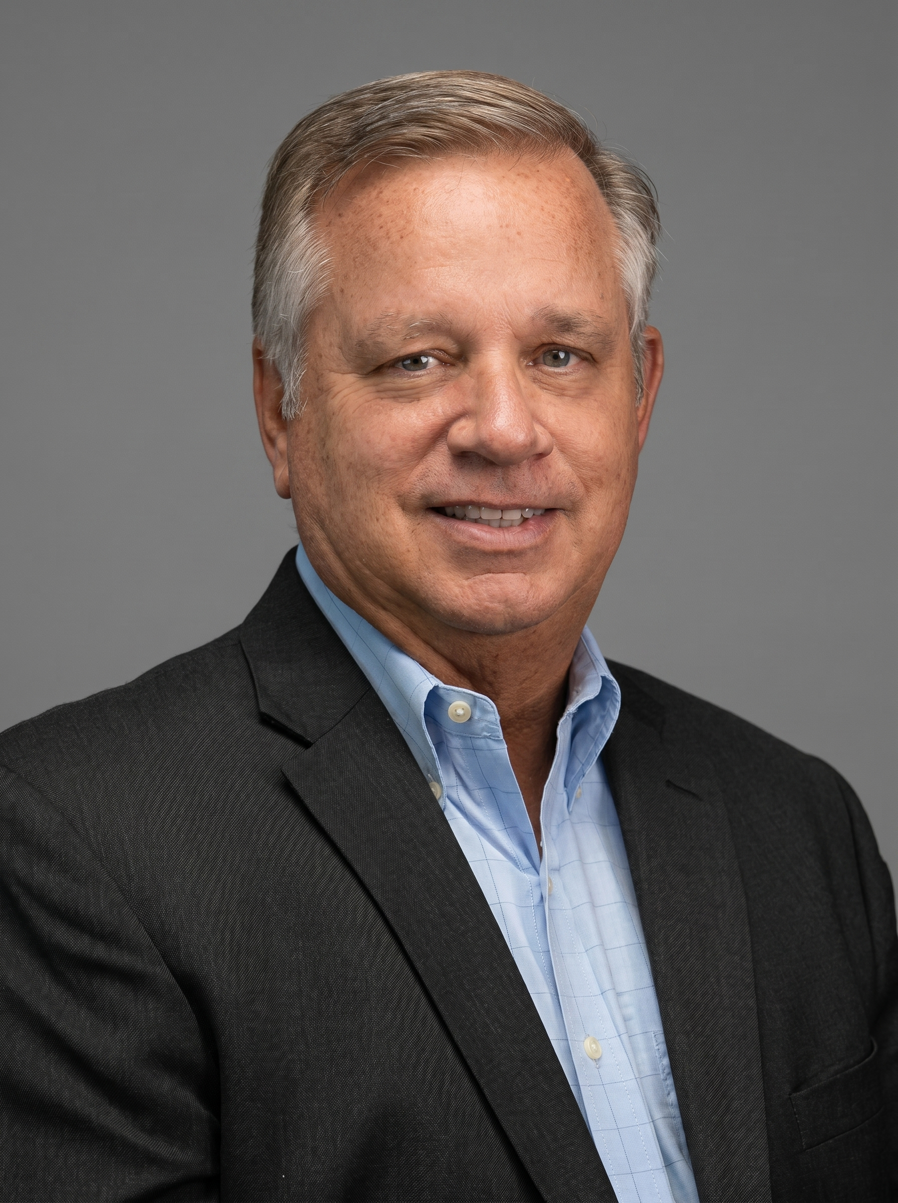Professional headshot of a middle-aged man wearing a dark suit jacket and a light blue dress shirt, against a plain gray background.