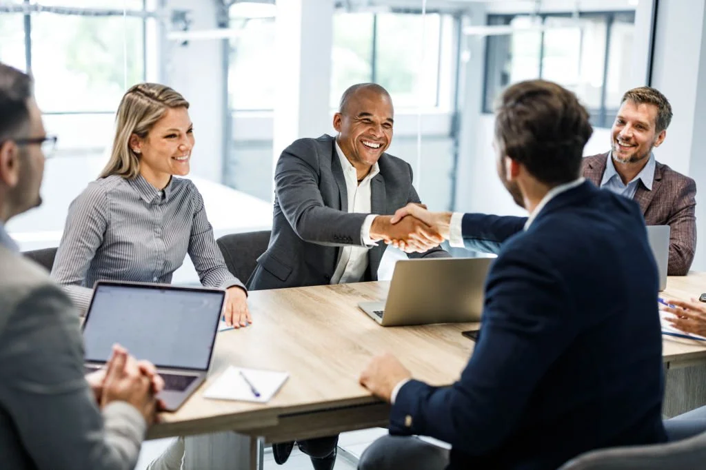 A diverse group of five professionals in business attire having a meeting in a modern office, with two people shaking hands across the table, and laptops and notepads in front of them.