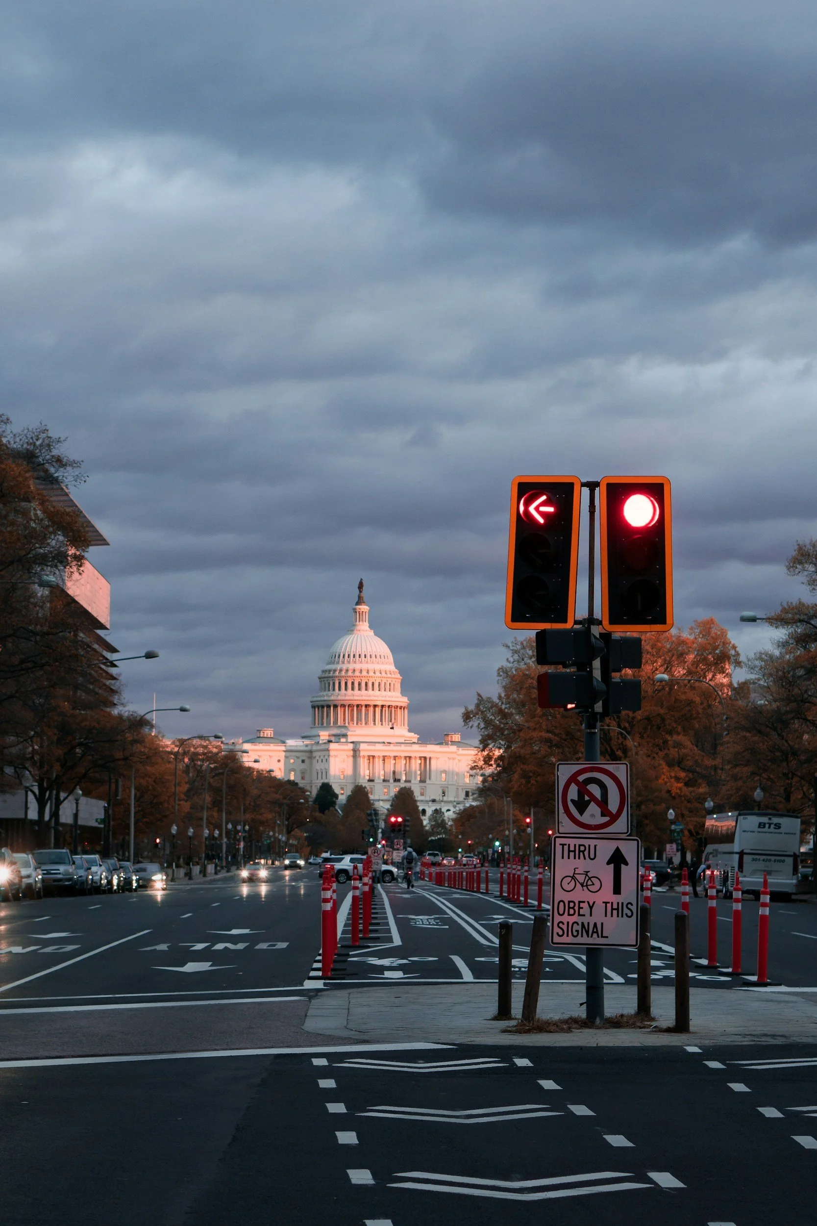 Street view of the United States Capitol building in Washington D.C. with cloudy sky, traffic lights, cars, and a sign instructing cyclists to obey this signal.