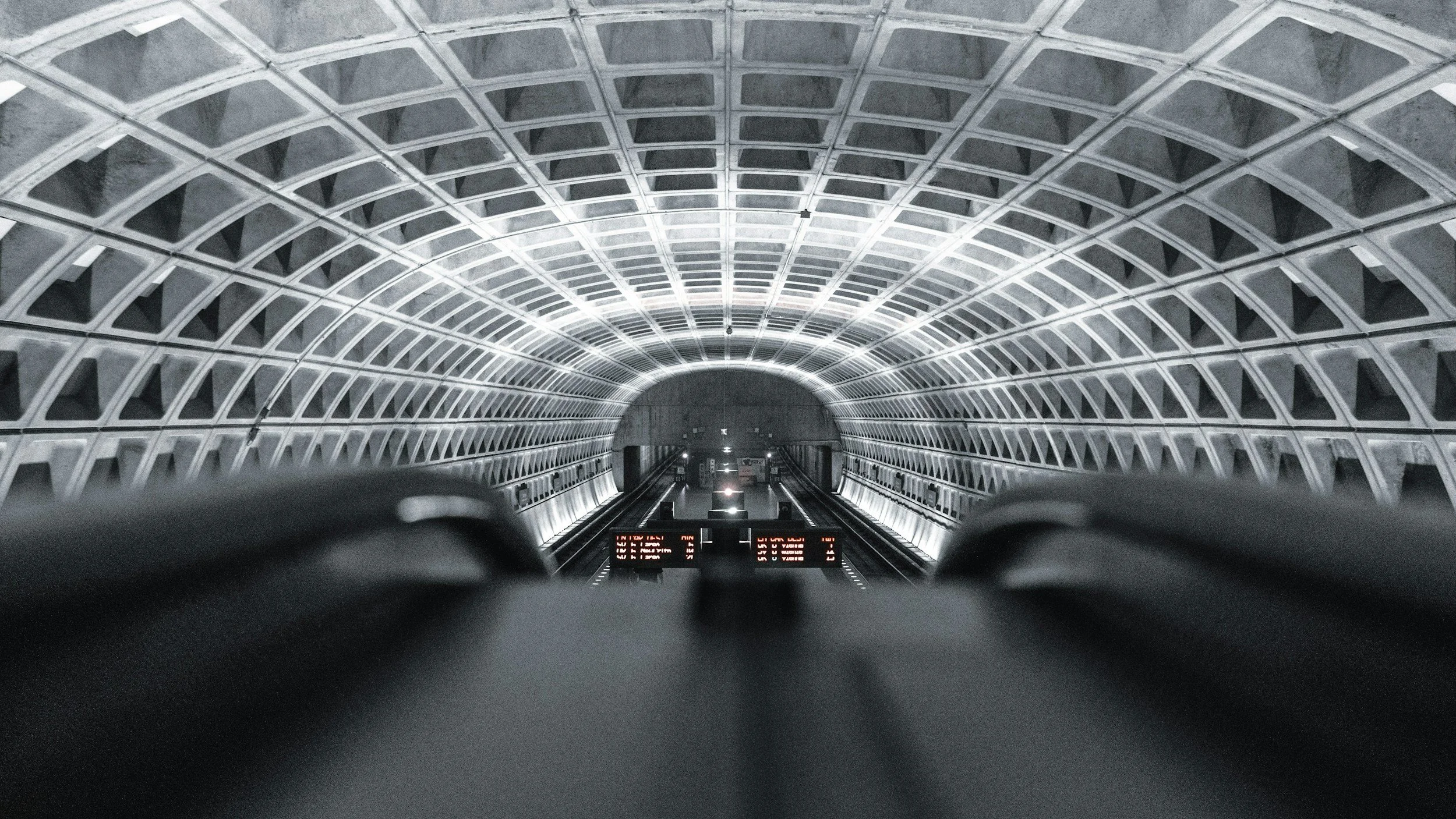 View from the bottom of an underground subway station escalator showing a curved, metallic ceiling with geometric patterns and LED strip lighting.
