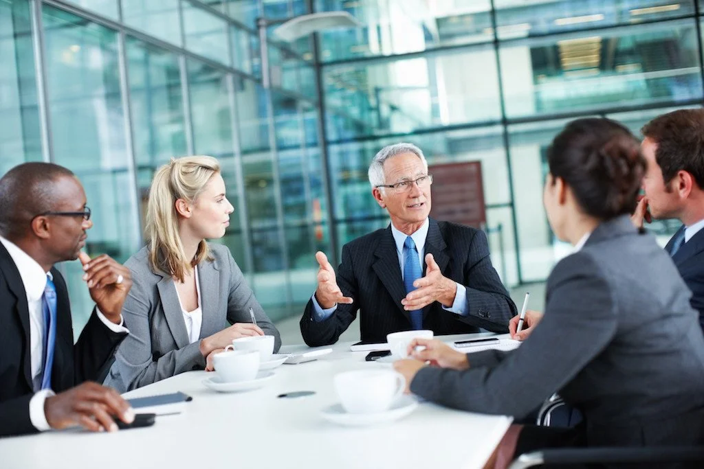 Business meeting in a modern office with five diverse professionals sitting around a table, engaging in a discussion.