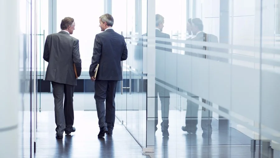 Two businessmen walking and talking in a modern office hallway with glass walls.