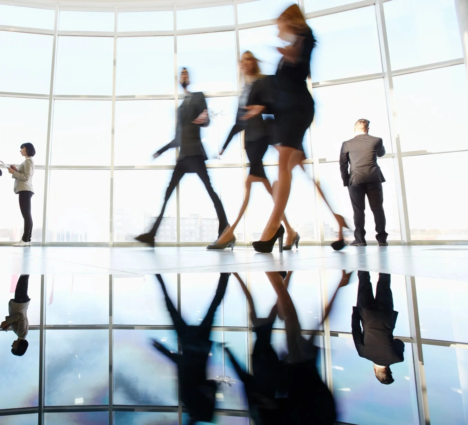 Businesspeople walking inside a building with large glass windows, reflecting some individuals and a bright sky outside.
