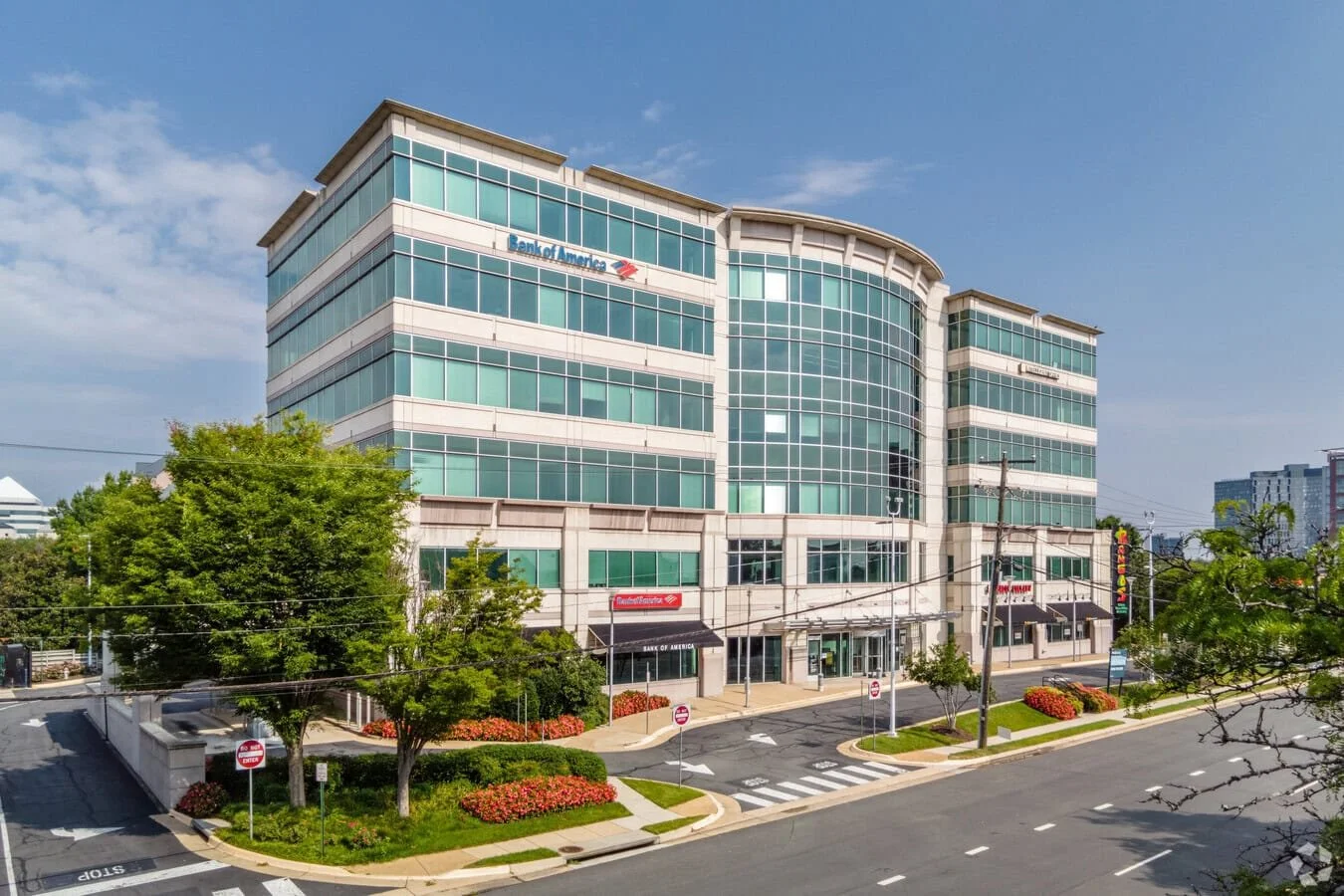 A modern multi-story office building with glass windows, featuring the Bank of America logo, situated on a street lined with trees, flowerbeds, and traffic signs.