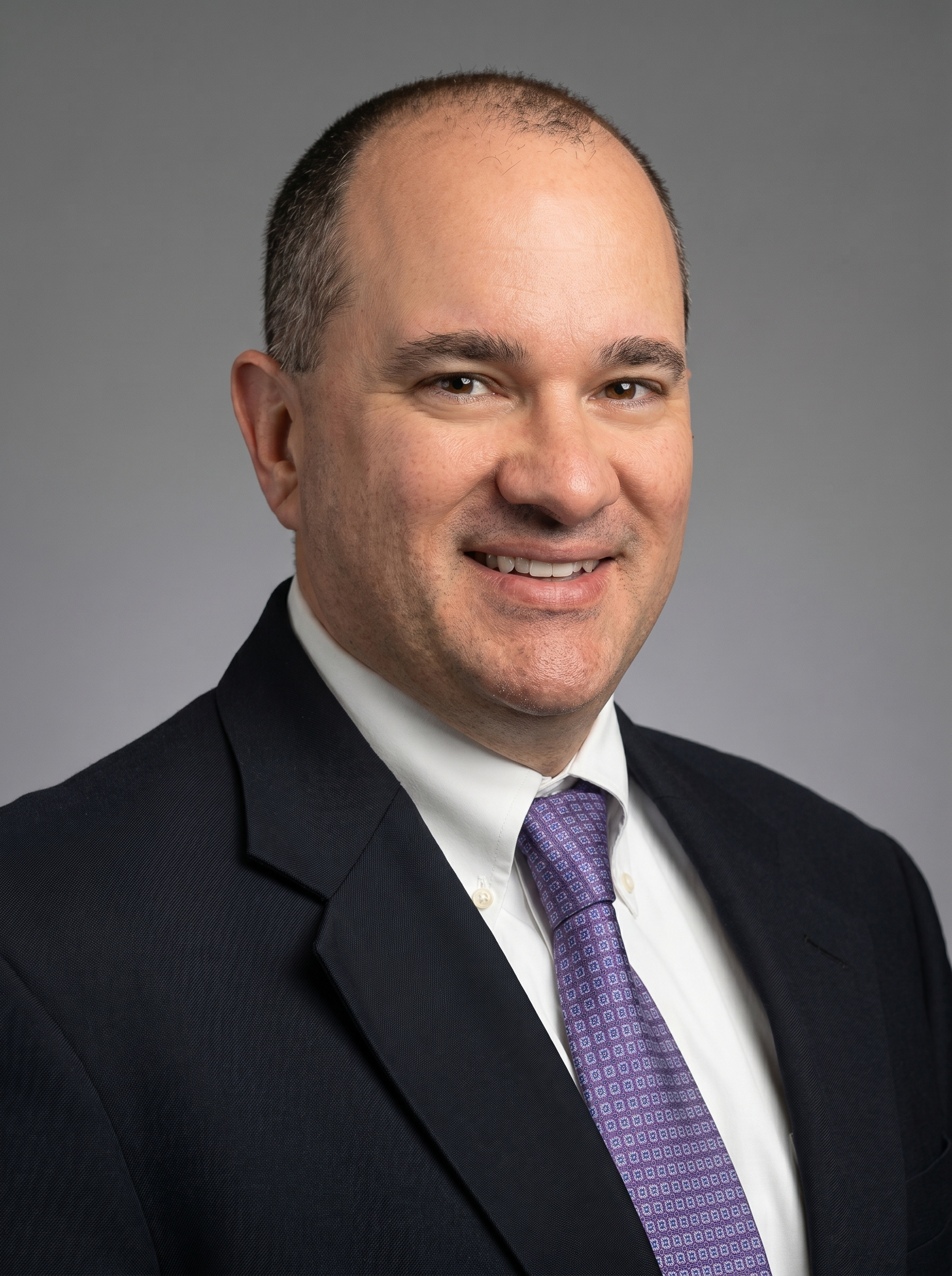 A professional headshot of a man in a dark suit, white shirt, and purple patterned tie, smiling against a gray background.