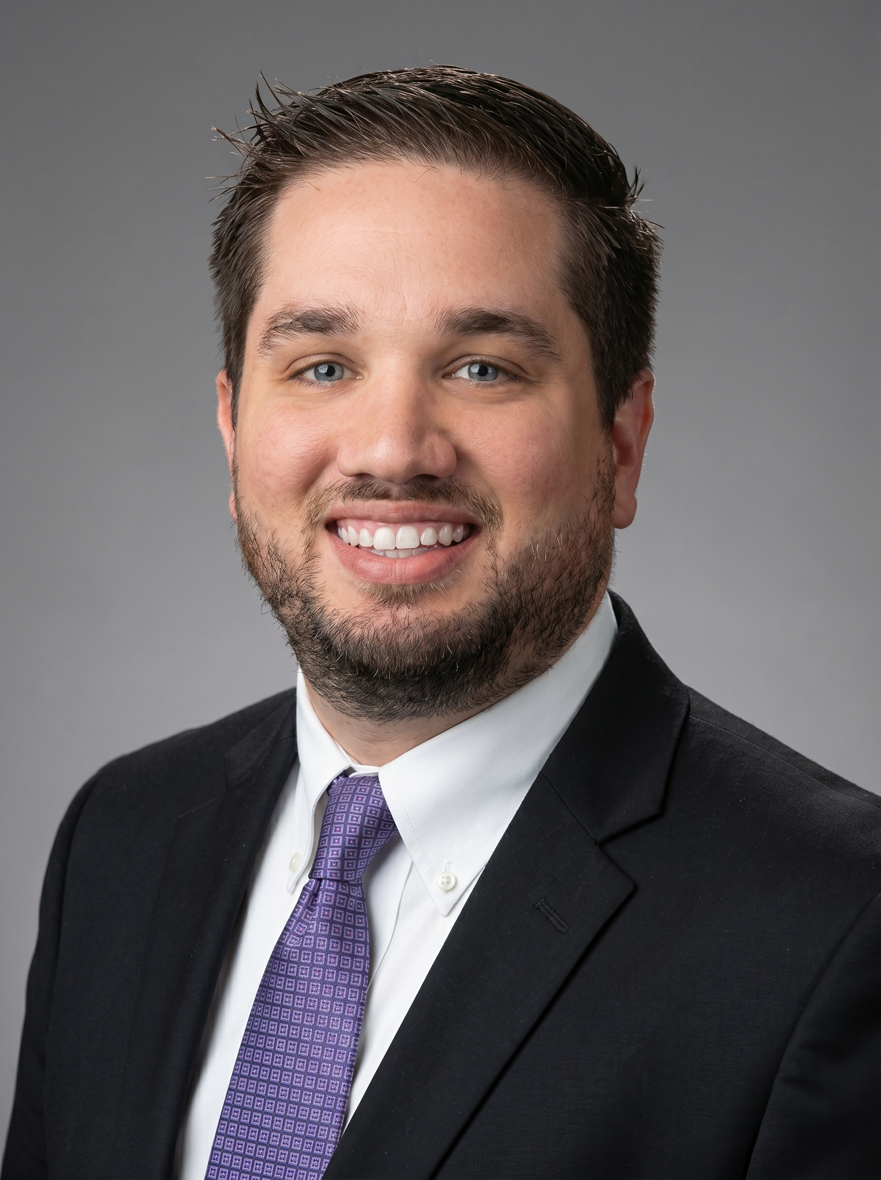 A smiling man in a suit, wearing a purple patterned tie, with dark hair styled upward, and a beard, against a gray background.