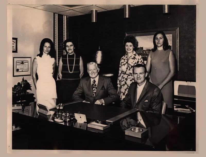 Six individuals in a professional office setting, three men and three women, dressed in formal attire, with papers and office supplies on the desk.