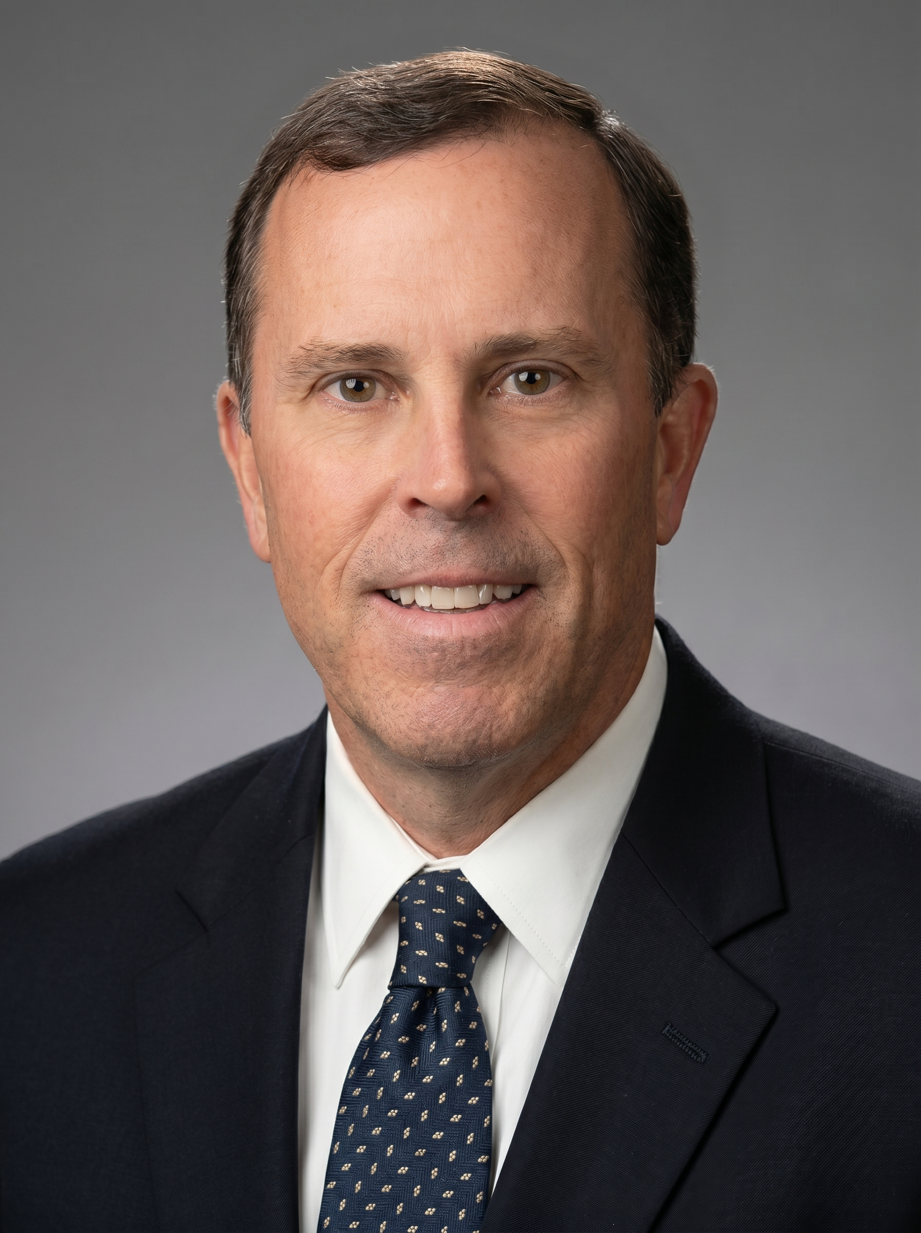 Close-up of a smiling man in formal business attire, with dark suit, white shirt, and patterned blue tie, against a gray background.