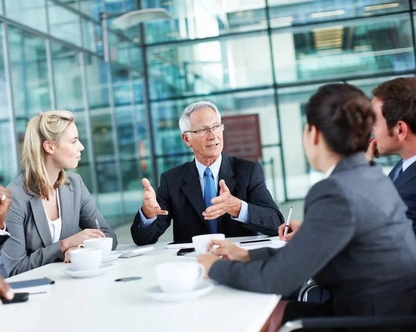 Business meeting with five people sitting around a table in a modern glass office, engaged in a discussion.
