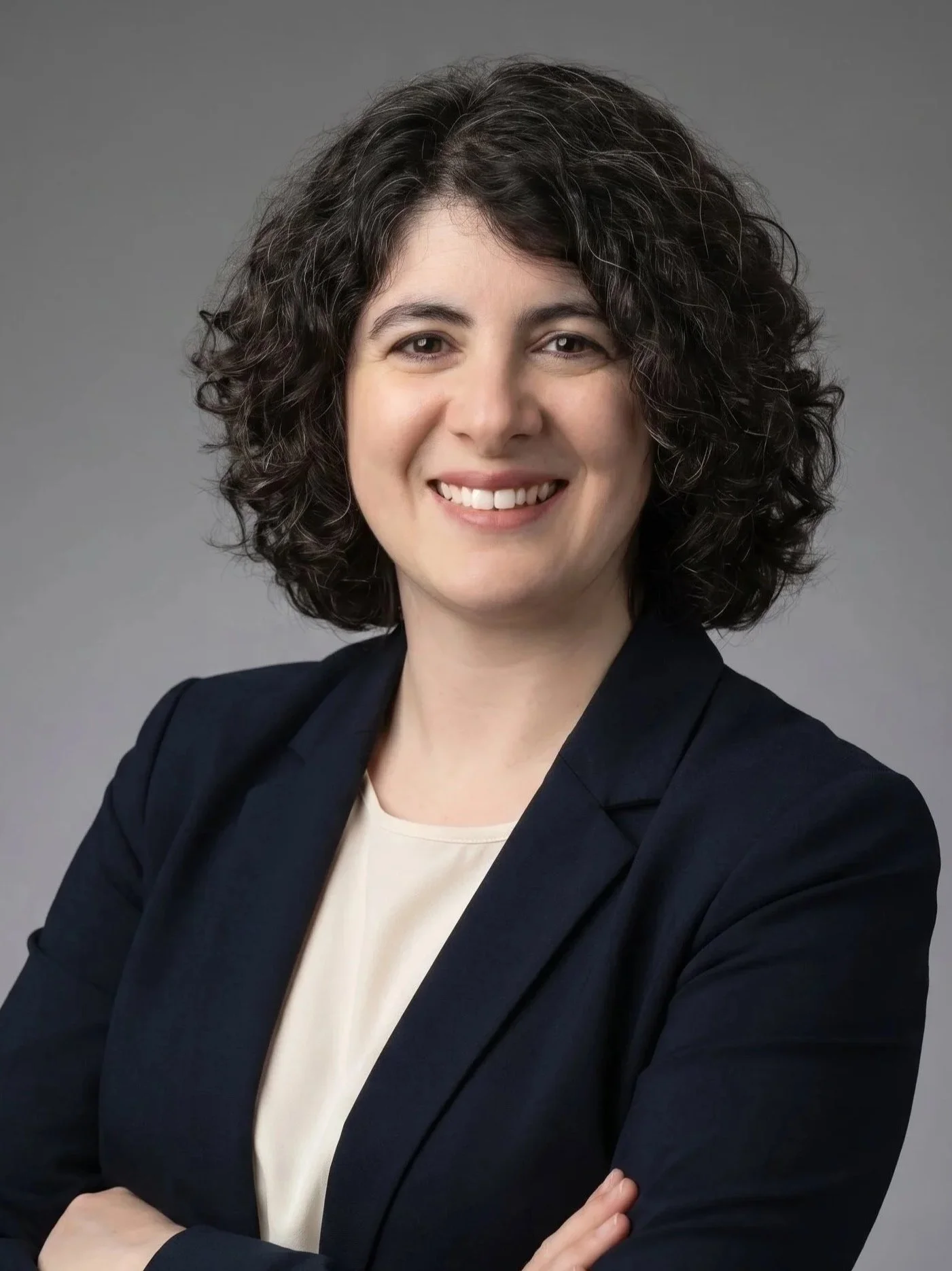 A woman with curly dark hair wearing a black blazer and a light-colored top, smiling and crossing her arms, against a gray background.
