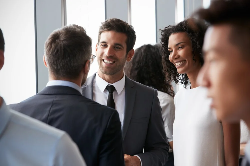 Group of diverse professional people chatting and smiling at a business event in a well-lit office or conference room.