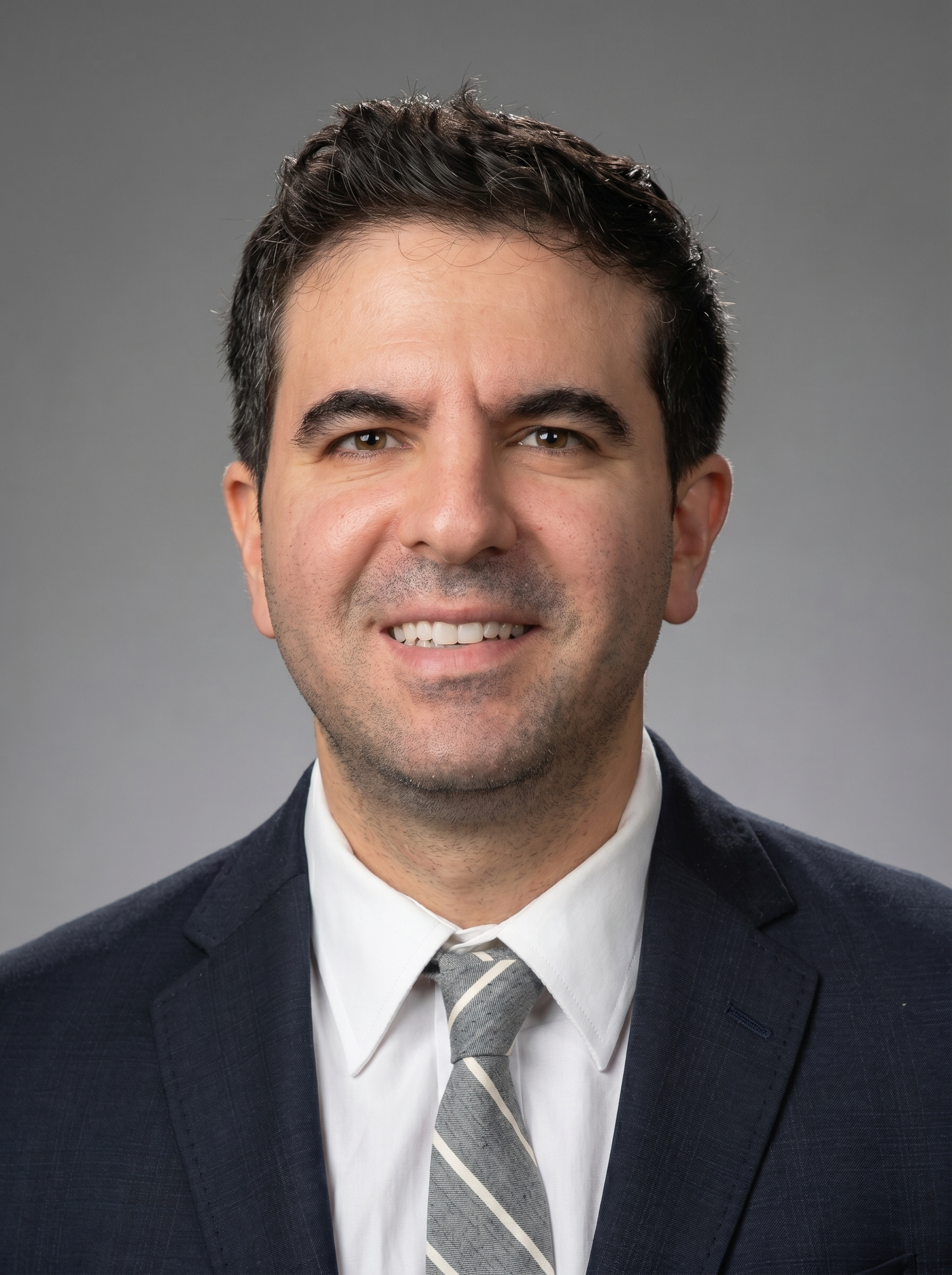 Headshot of a smiling man in a formal suit with a striped tie, light skin, dark hair, and brown eyes against a gray background.