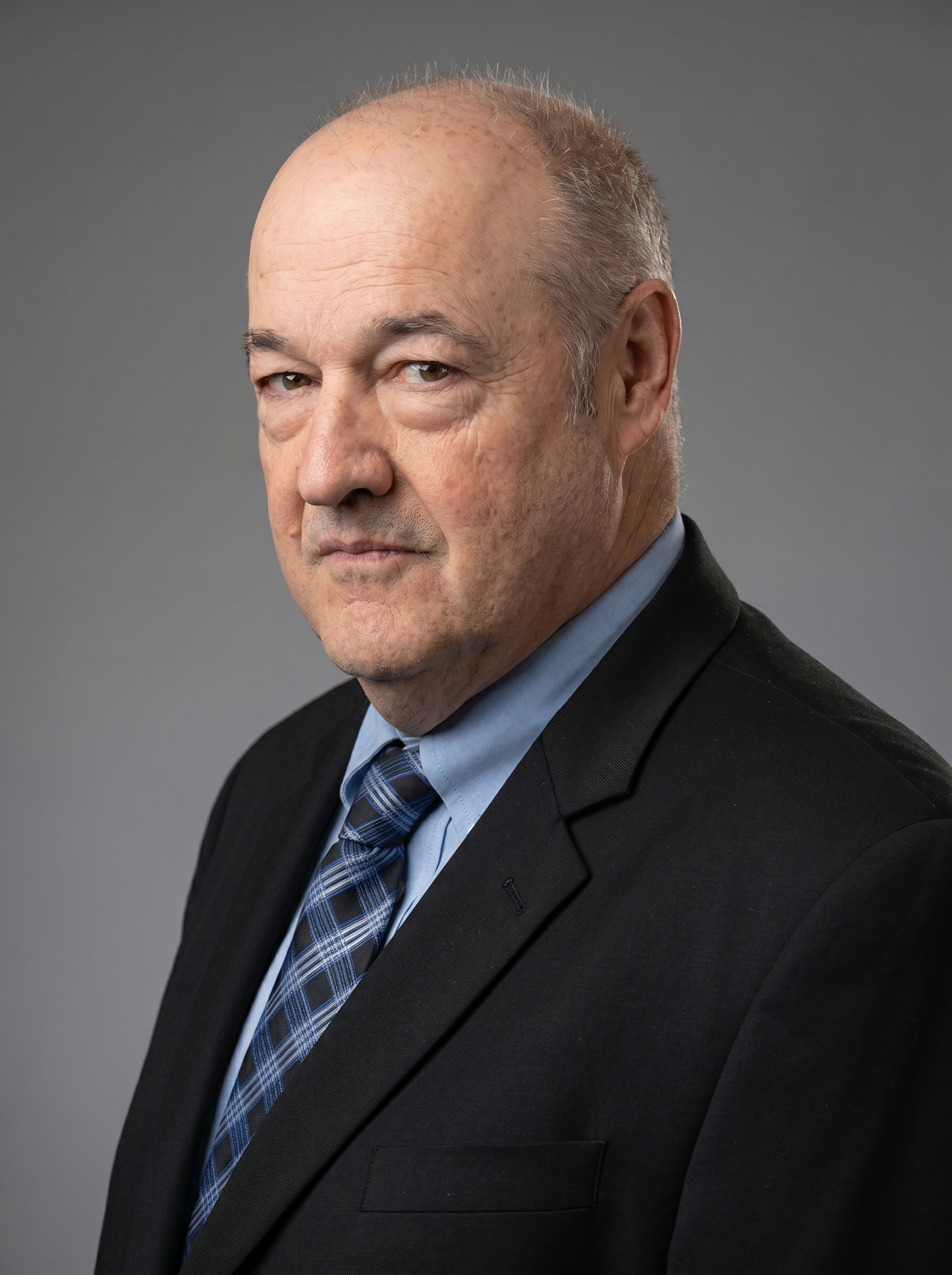 Portrait of a middle-aged man with a serious expression, wearing a black suit, light blue shirt, and a blue plaid tie, against a plain gray background.
