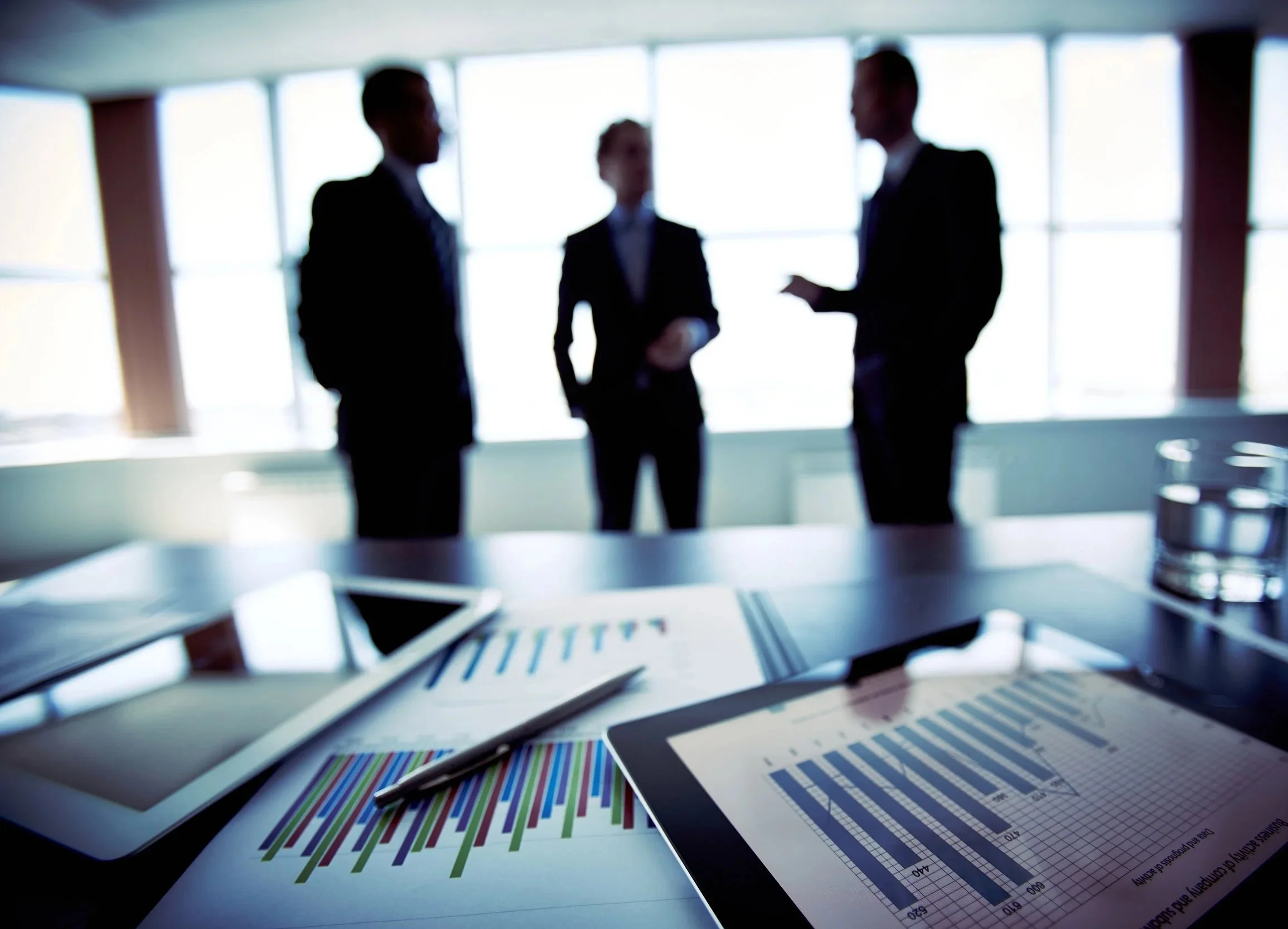 Close-up of financial documents and a pen on a desk with a laptop, with a blurred background of three business professionals having a discussion in a modern office with large windows.