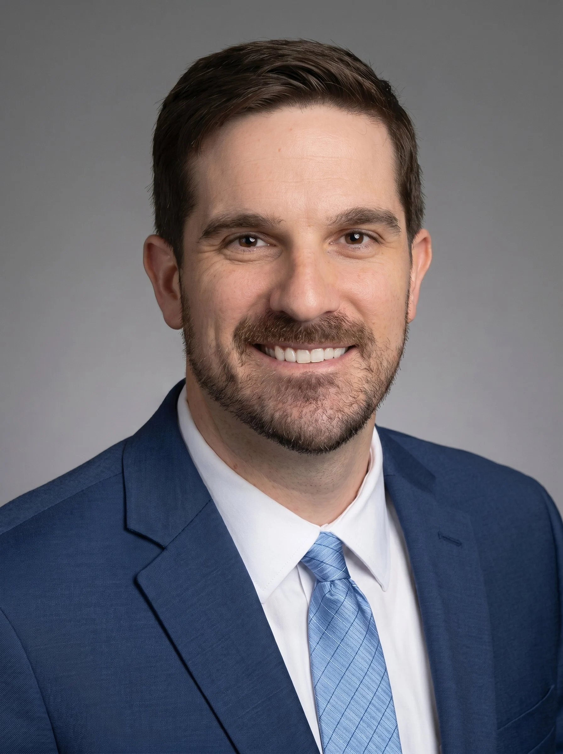 Portrait of a man in a blue suit and tie, smiling with visible teeth, short dark hair, beard, and mustache, against a plain grey background.