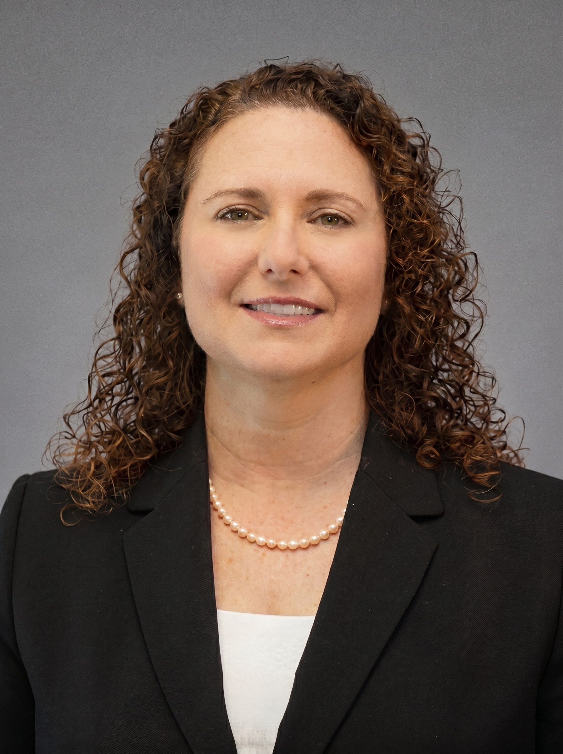 Portrait of Meredith Yenis, an executive recruiter in finance, wearing a black blazer, white top, pearl necklace, and small pearl earrings, against a neutral gray background.