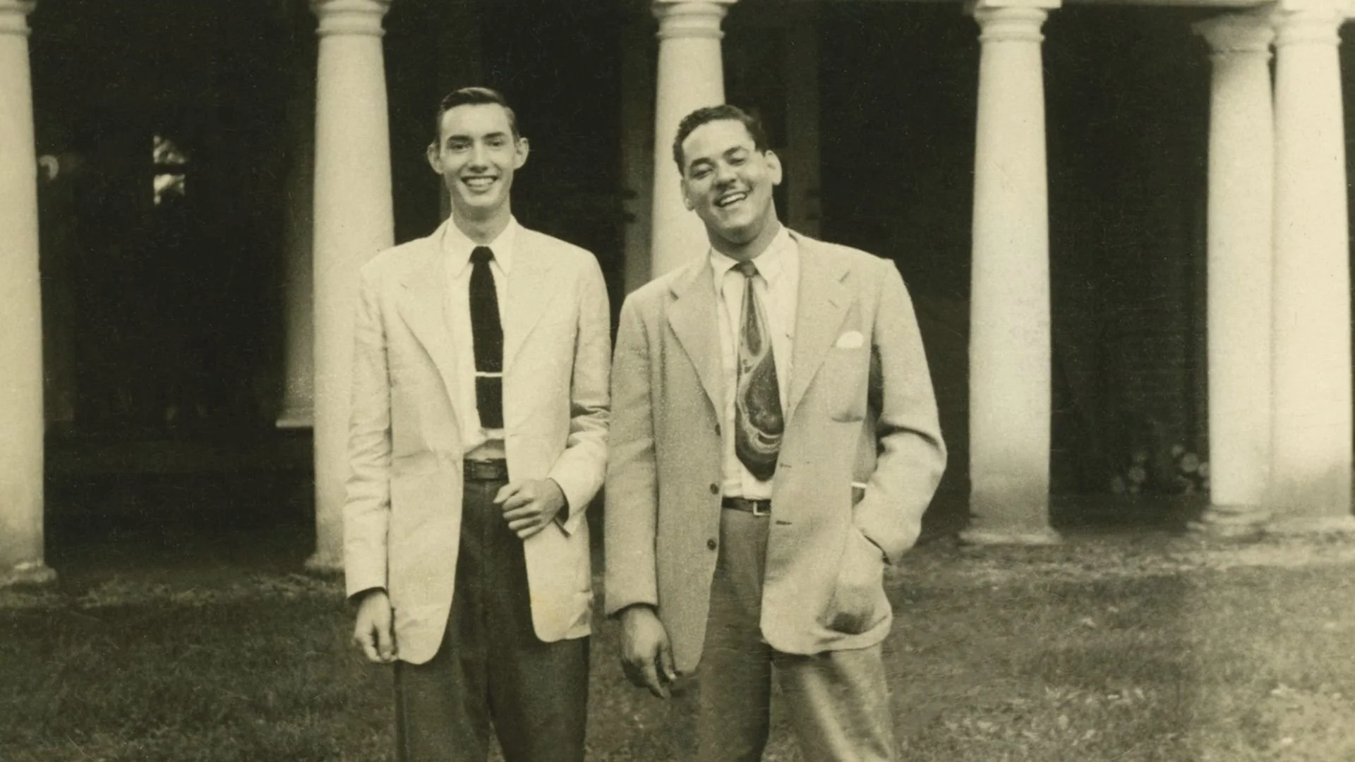 Two young men wearing suits and ties stand in front of a row of white columns on a grassy area, smiling at the camera in a black and white photo.