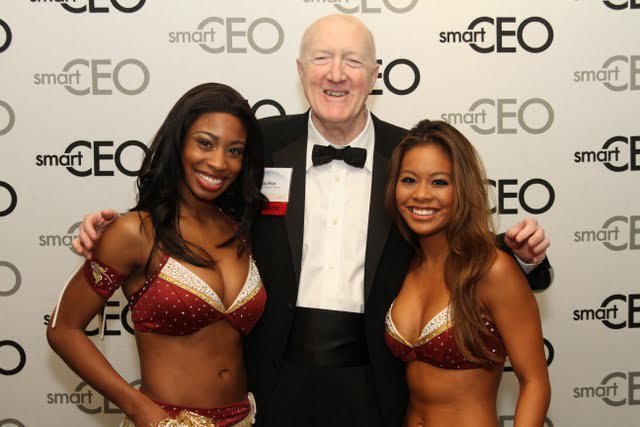 Two women in bikinis standing with an older man in a tuxedo at a smartEO event