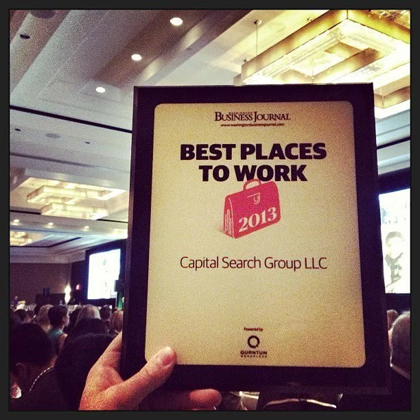 A person holding up an award certificate at a conference, titled 'Best Places to Work 2013', awarded to Capital Search Group LLC, during a Business Journal event in a large conference room filled with attendees.